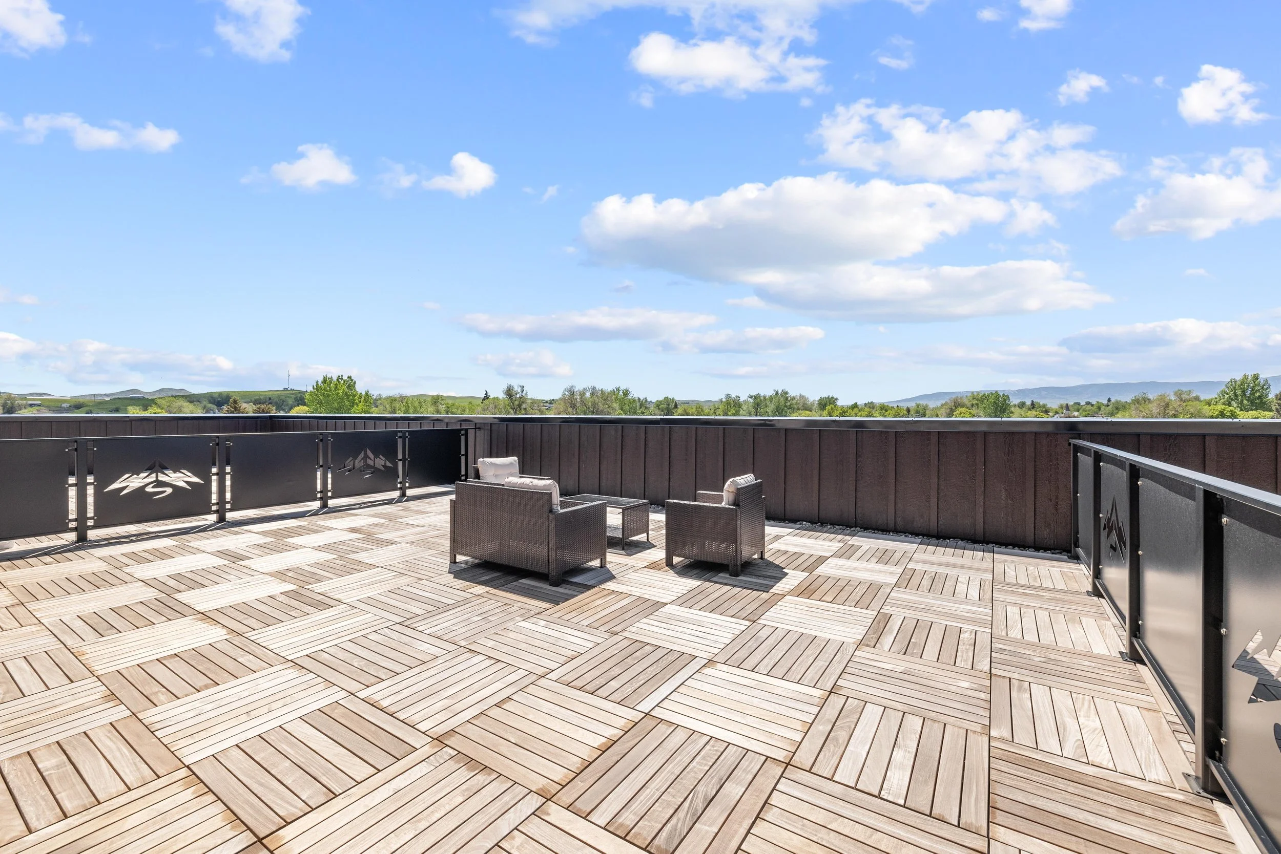 Large outdoor rooftop deck with wooden tile flooring, black metal railing with mountain logo cutouts, some patio furniture including chairs and small table, overlooking green landscape and cloudy sky.