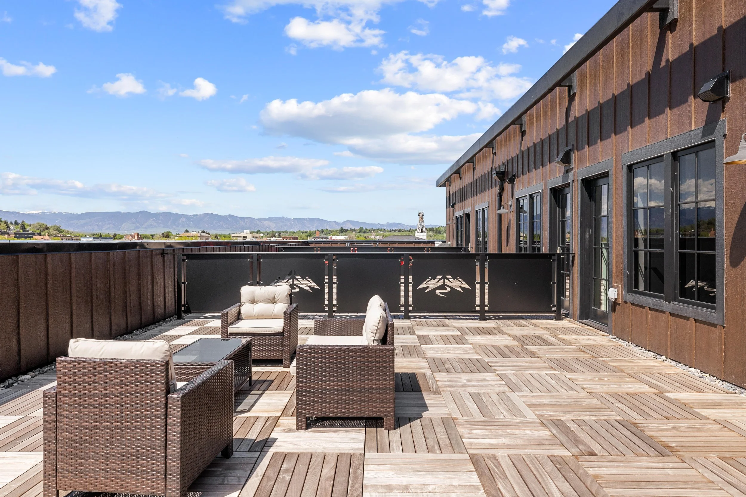 Outdoor rooftop patio with wicker chairs and glass table, wooden deck flooring, black metal railing, brown exterior building wall, and mountain view in the background under a partly cloudy sky.