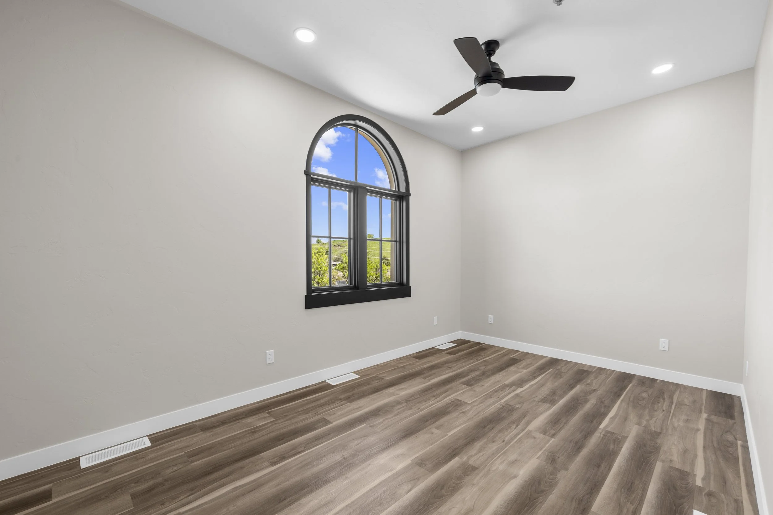 Empty room with beige walls, wood flooring, black-framed arched window, ceiling fan, and recessed lighting.