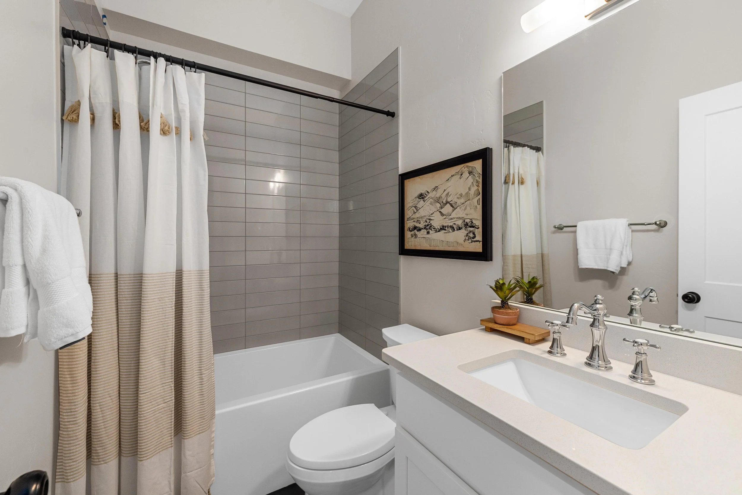 Bathroom with white and beige shower curtain, gray tiled shower area, white sink with chrome faucet, mirror, towel rack with white towel, wall art, and small plants on the counter in an Ice Haus Condo for sale in Sheridan, Wyoming 