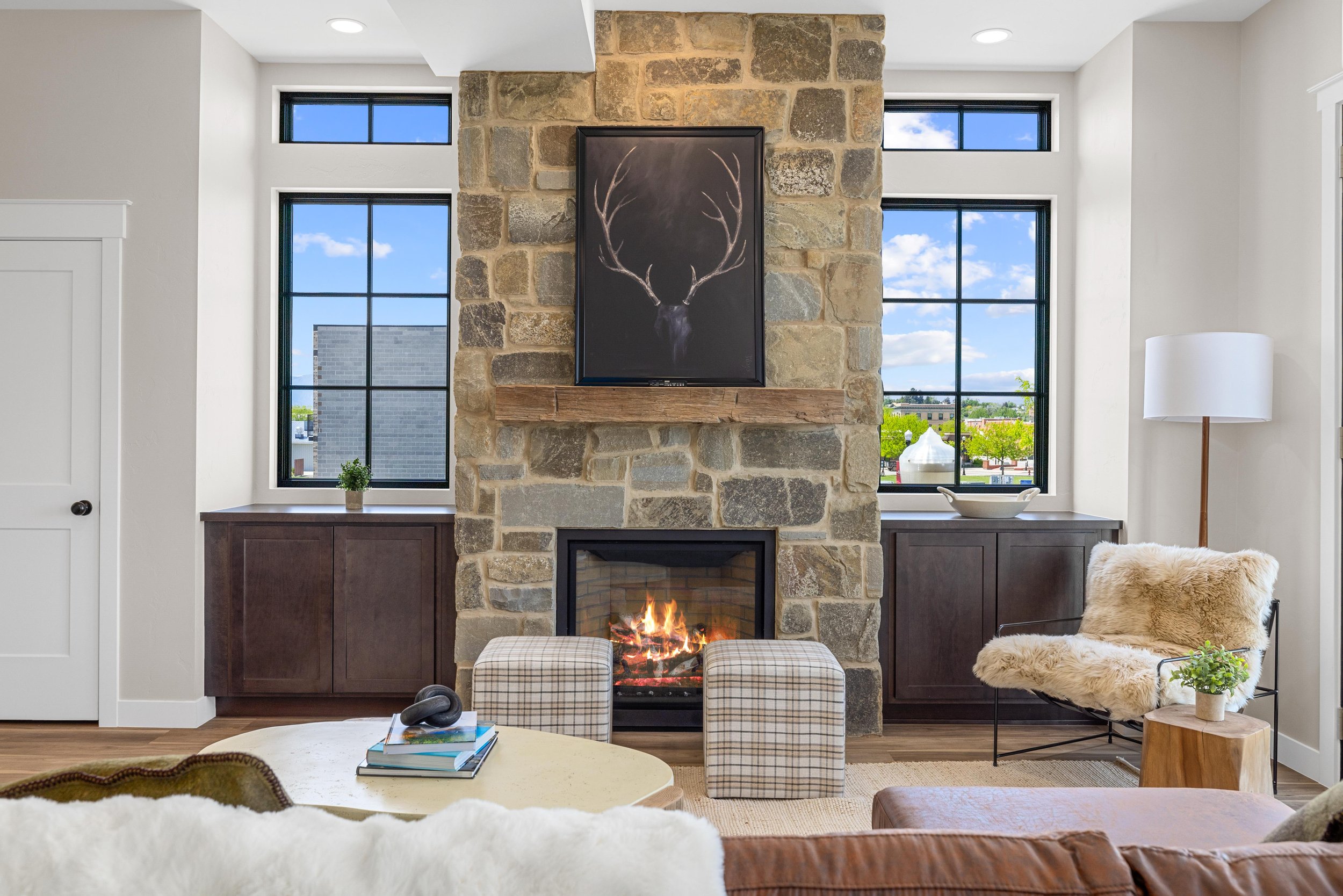 Living room with a stone fireplace, two large black-framed windows, a wooden mantel with a framed antler artwork, a cozy chair with a faux fur throw, a white floor lamp, a wooden side table with a plant, and a beige ottoman with a pair of headphones 