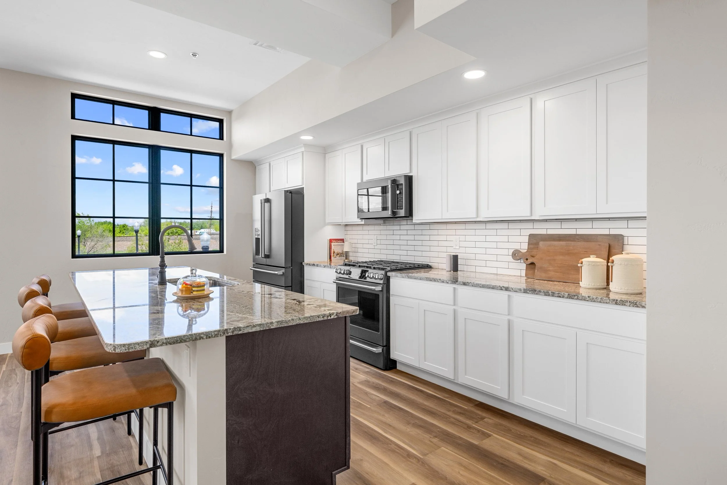 Modern kitchen with white cabinets, gray countertops, black appliances, wooden flooring, and a large window with a view of green trees and blue sky.