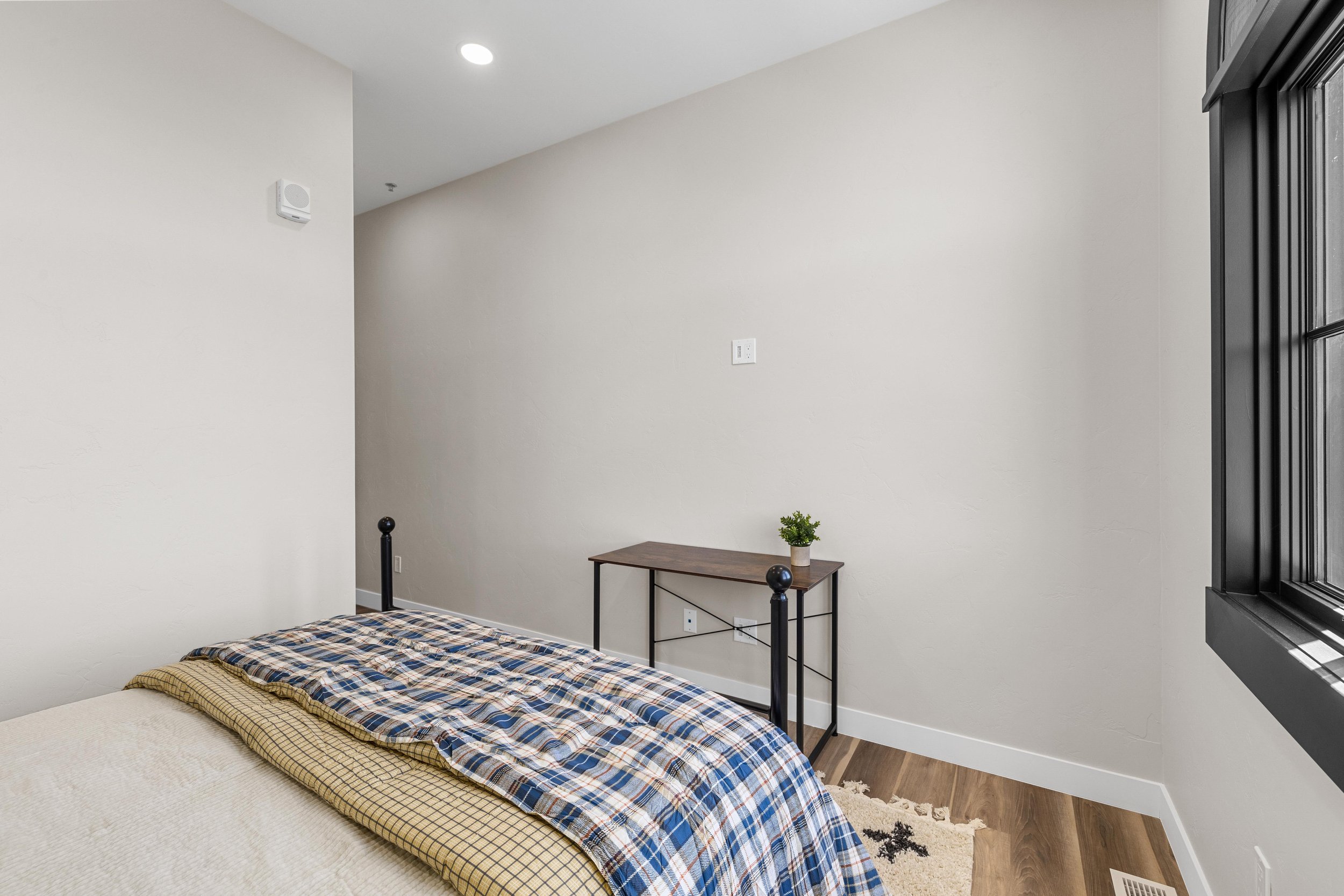 A bedroom corner with a bed covered in a plaid blanket, a small wooden table with a potted plant, and a window with black trim, in an Ice Haus Condo for sale in Sheridan, Wyoming.