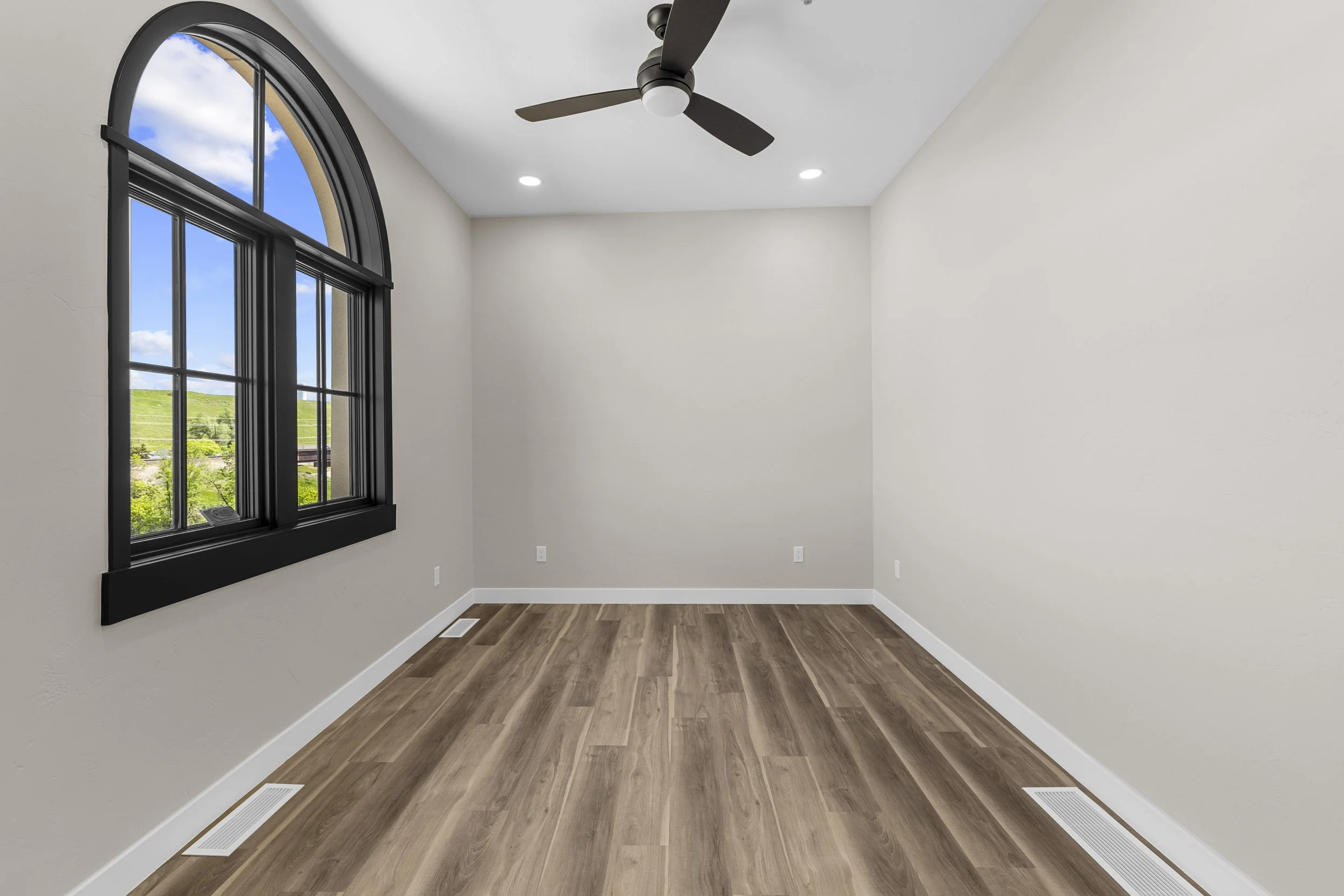 Empty room with beige walls, a large arched window with black trim, a ceiling fan, wood flooring, and natural light.