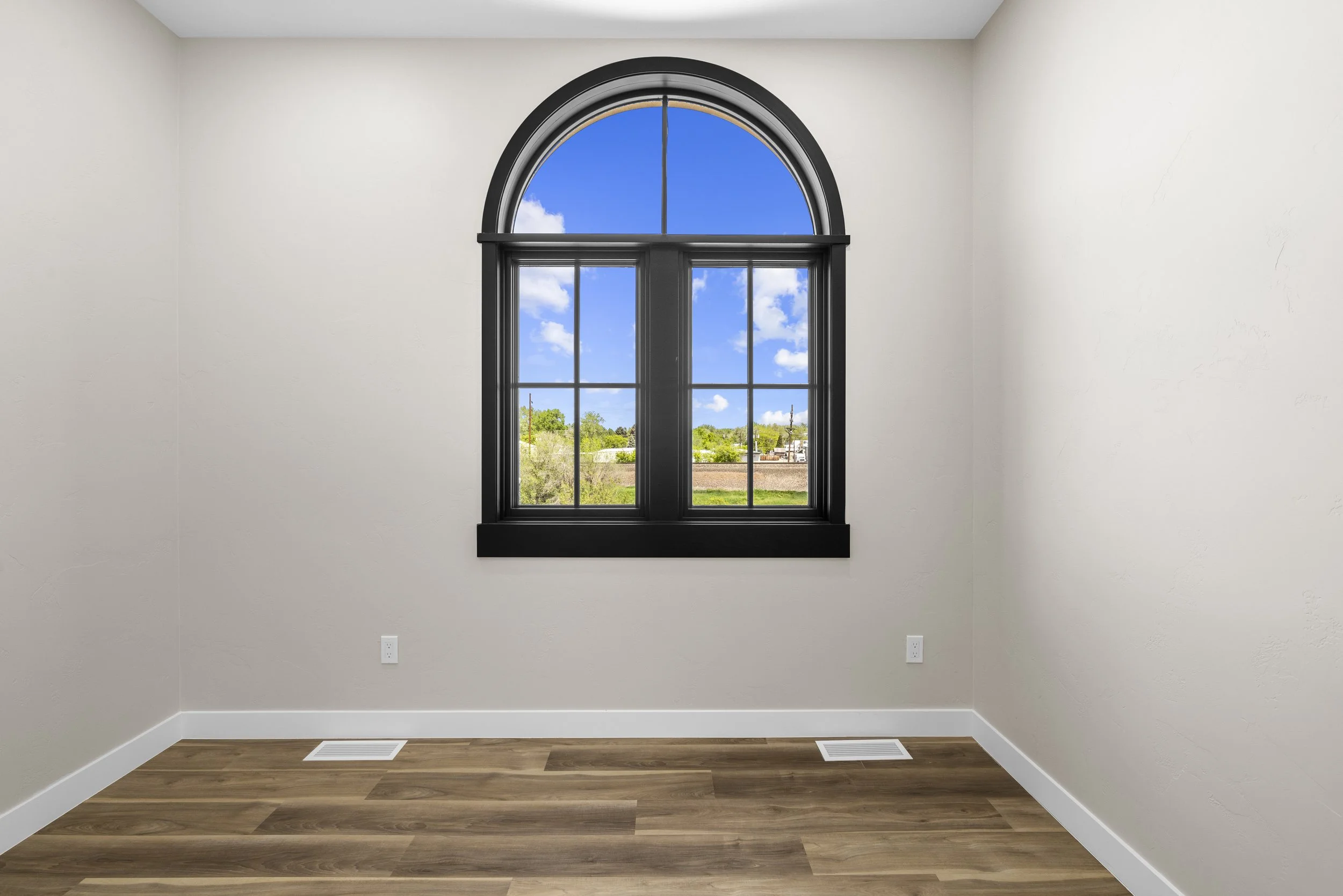 Empty room with beige walls, hardwood floor, black-trimmed window, and blue sky with clouds outside.