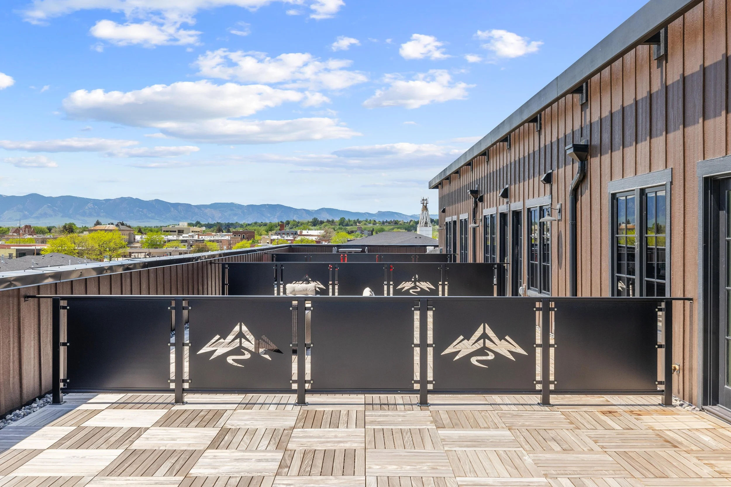 Outdoor balcony with wooden tiles, black metal privacy screens with mountain logo, brown siding, multiple windows, and a scenic mountain view in the distance.