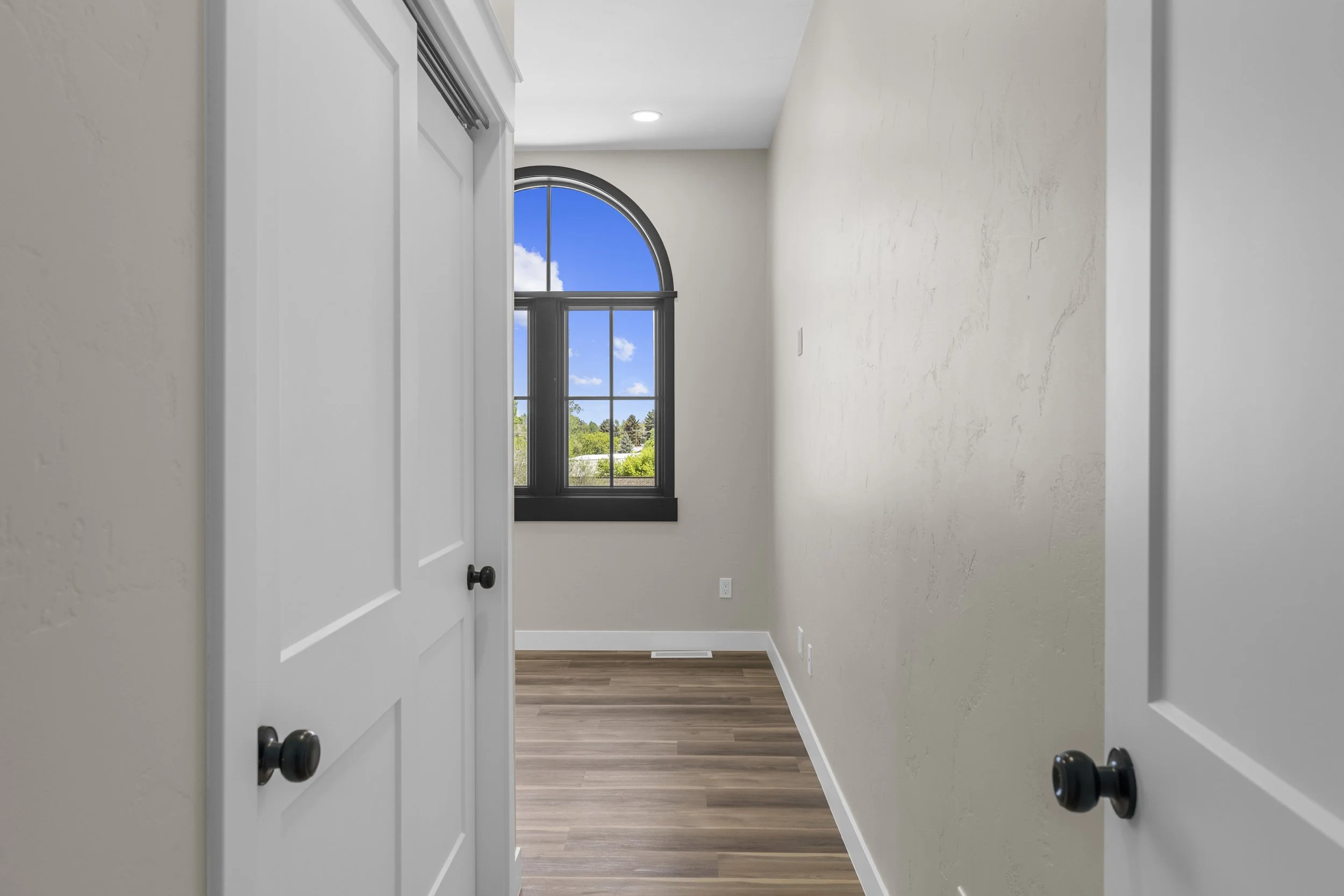 Empty room with white walls, a wooden floor, a black arched-framed window showing a blue sky with clouds, and white closet doors on the left.
