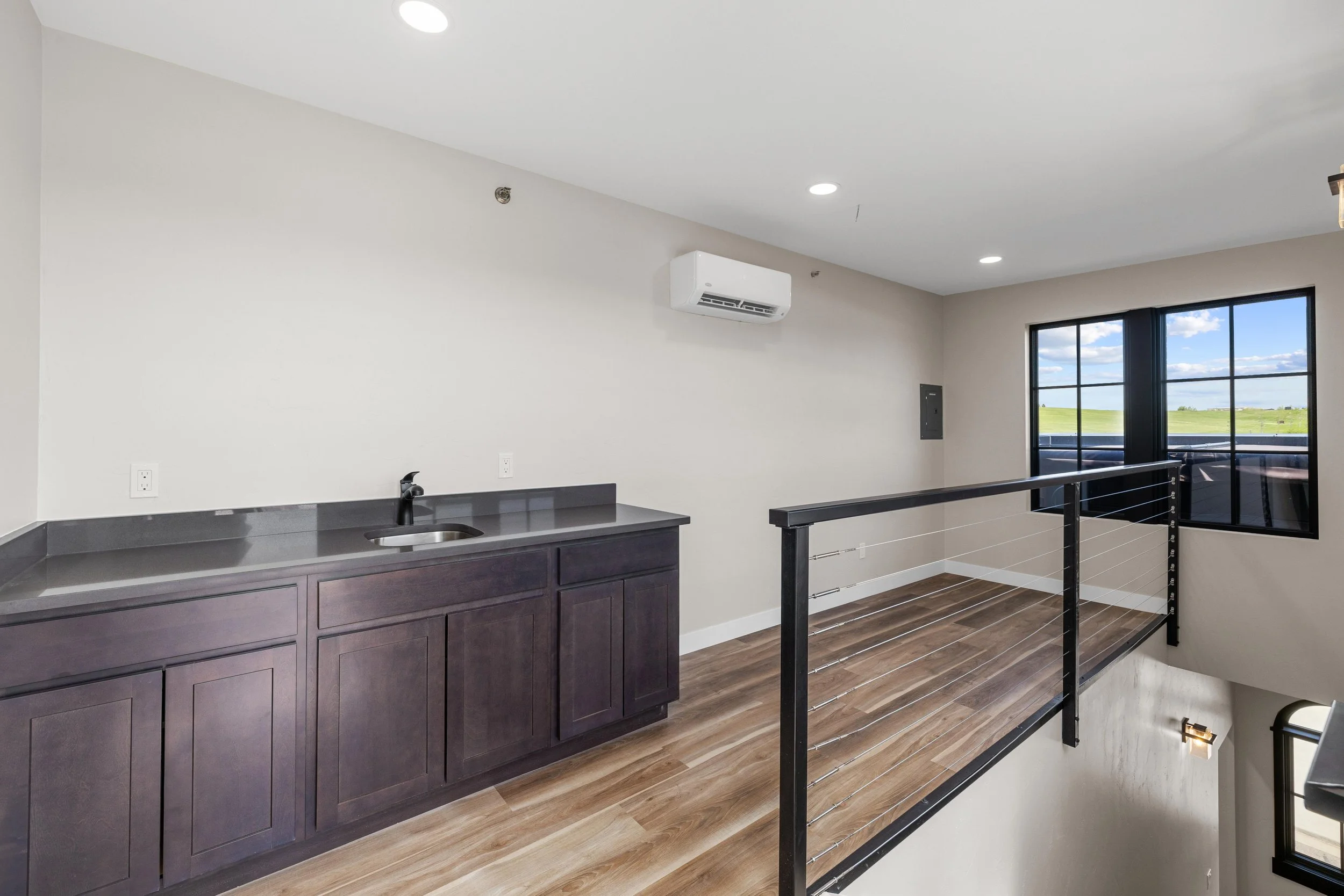 Interior view of a modern room with a dark wood kitchenette, black countertop with a sink, large window showing a green landscape, and black metal railing along a small open loft space with wood flooring.