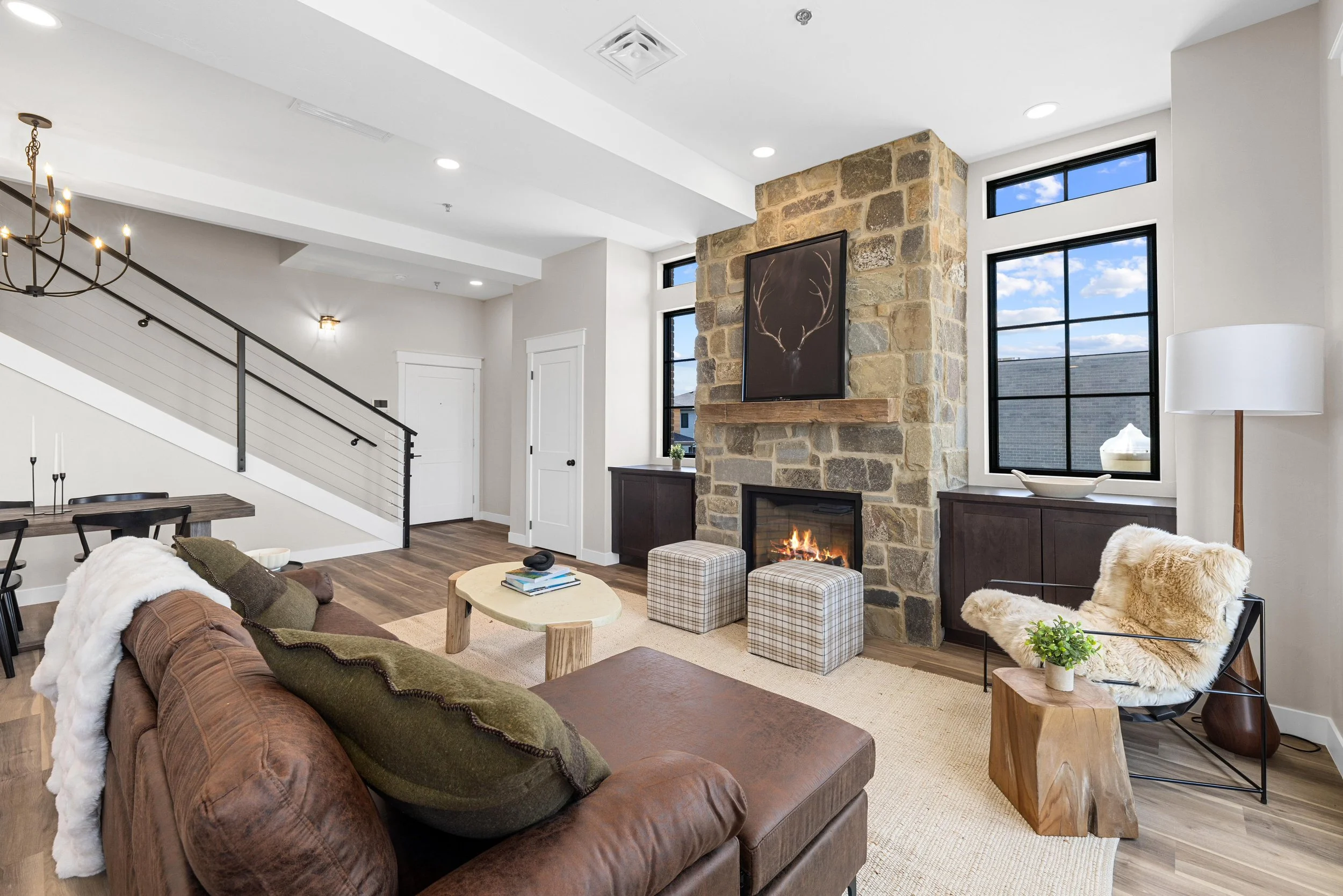 Living room with stone fireplace, large black-framed windows, brown leather sofa with green and white pillows, modern chair with sheepskin cover, wooden side table with plant, plaid ottomans, and a beige rug.