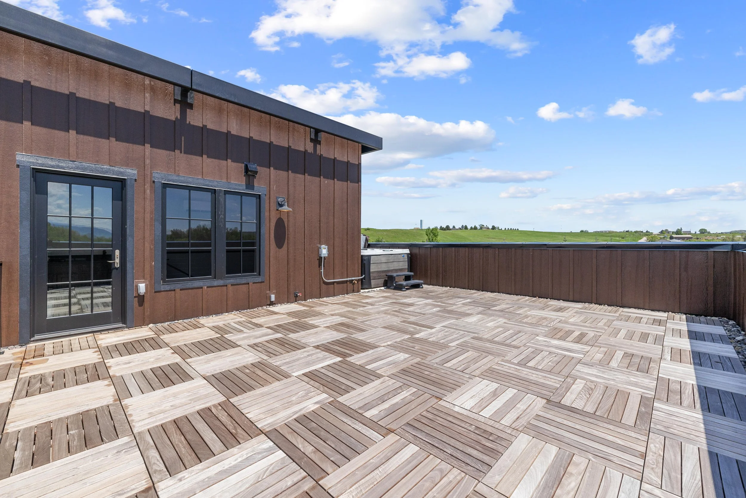 Rooftop terrace with wood tile flooring, brown siding wall with a door and windows, railing, and a view of green fields and blue sky with clouds.