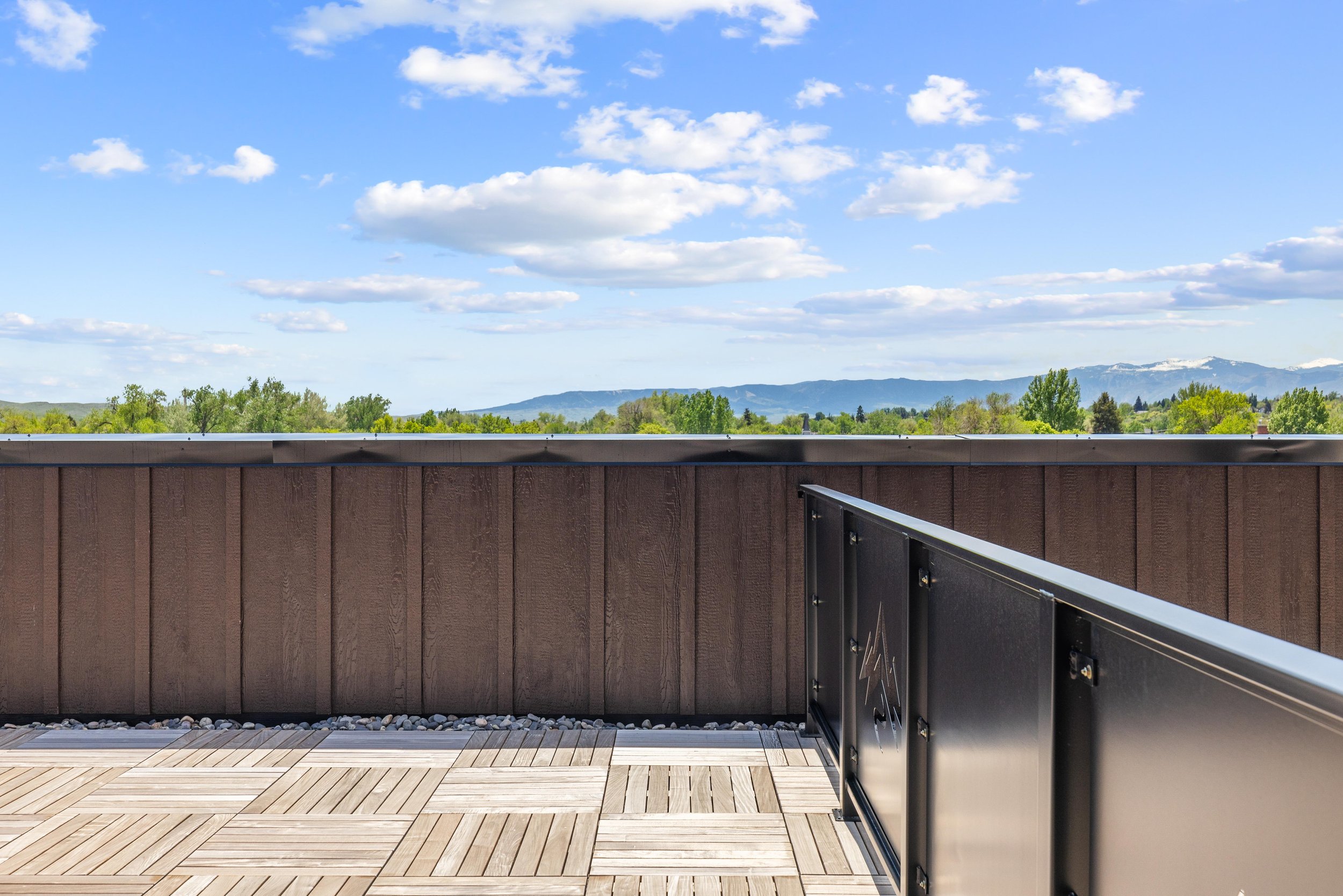 Rooftop balcony with wooden flooring and a brown wooden wall, overlooking distant mountains and a green landscape under a partly cloudy blue sky.