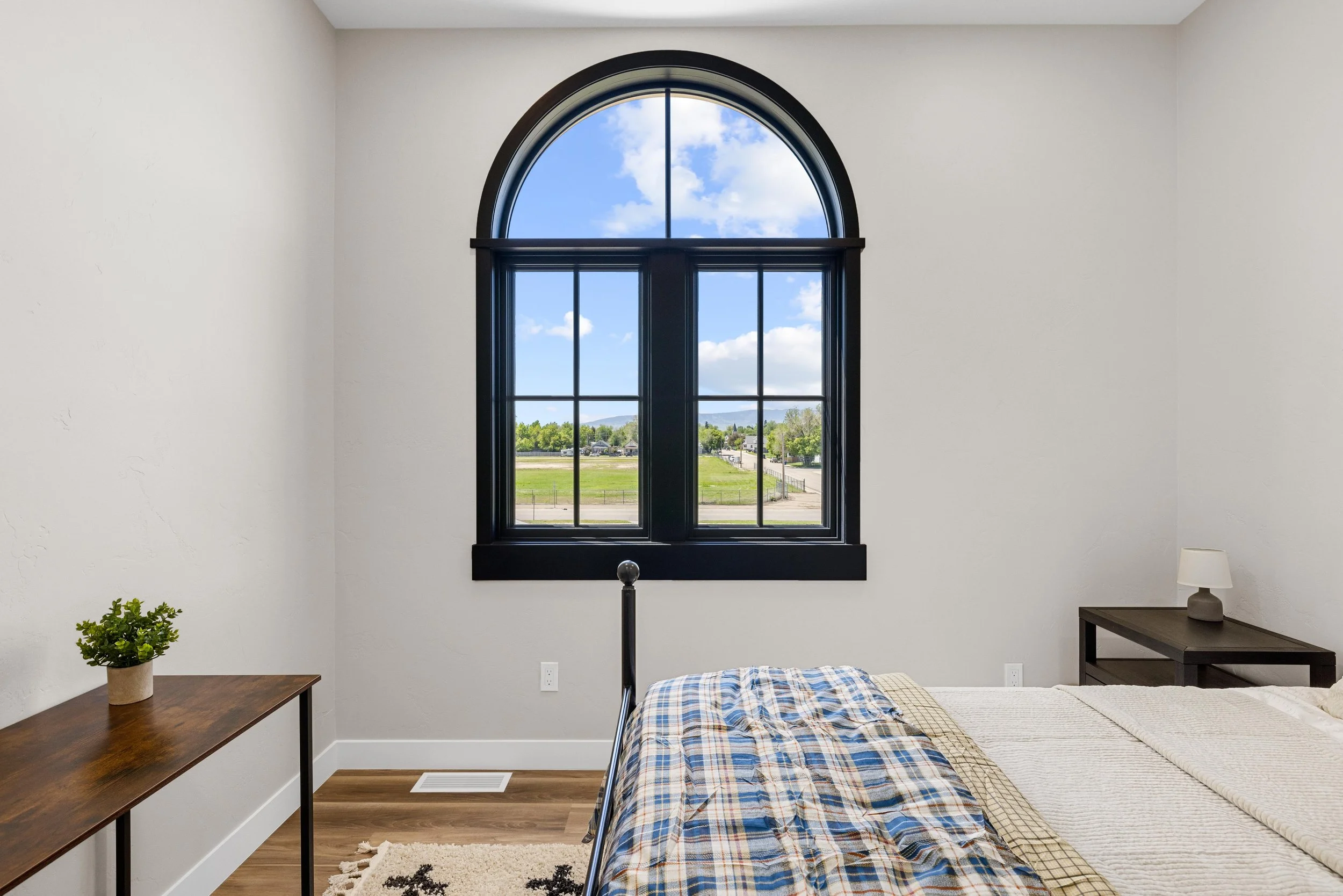 A bedroom with a large arched window showing a blue sky with white clouds and a green field outside. The room has a bed with a plaid blanket, a small table with a lamp on it, and a wooden desk with a potted plant, in an Ice Haus Condo for sale.