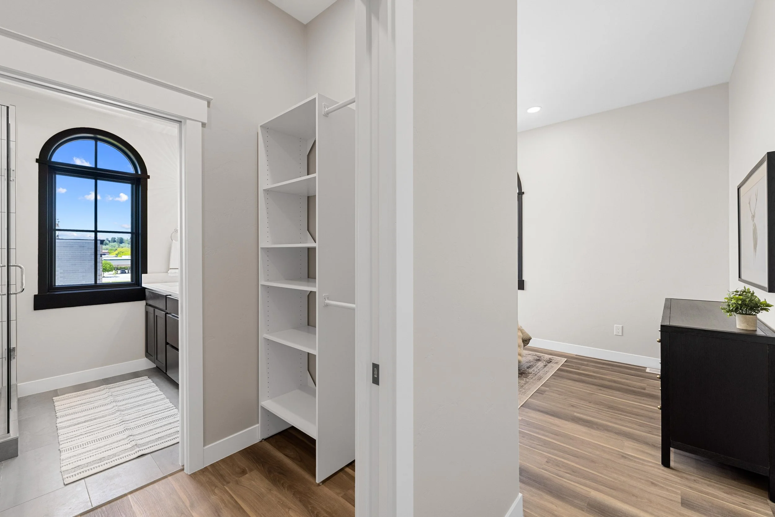Interior view of a home with a laundry room on the left featuring a black arched window, cabinets, and a striped rug, and a living area on the right with a black dresser, wall art, and wood flooring.