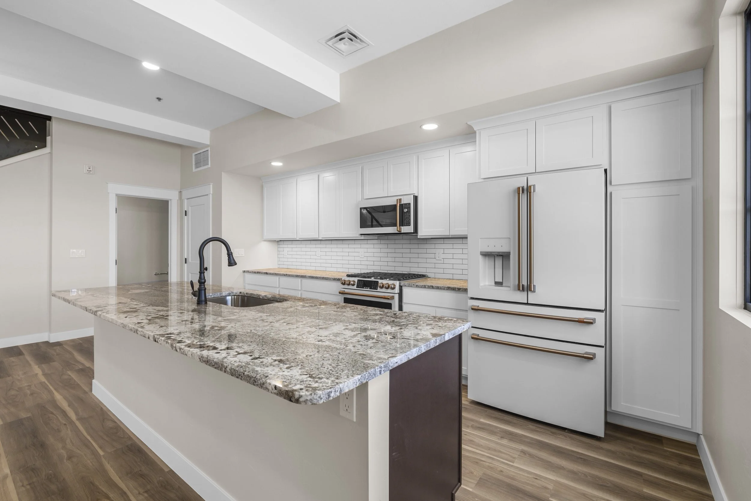 Modern kitchen with white cabinets, granite countertops, a black faucet, and stainless-steel appliances including a refrigerator, microwave, and oven, with wood flooring and a large window.