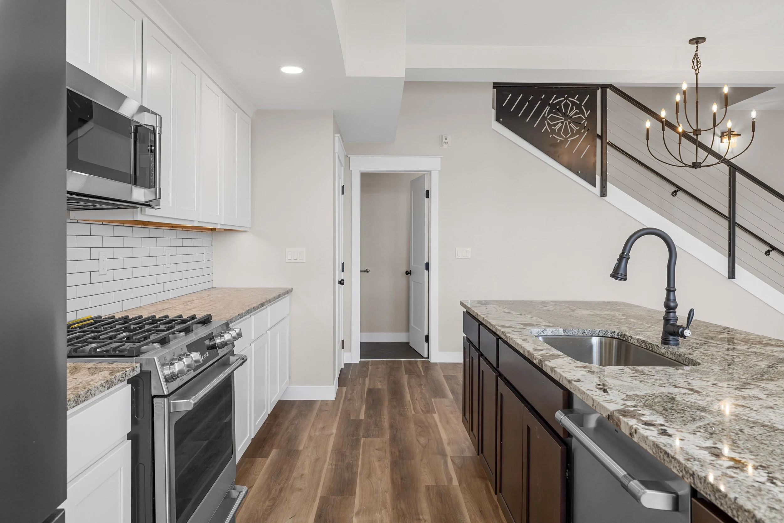 Modern kitchen with white upper cabinets, dark lower cabinets, granite countertops, stainless steel appliances, white subway tile backsplash, wood flooring, and a staircase with black railing and a chandelier in the background.