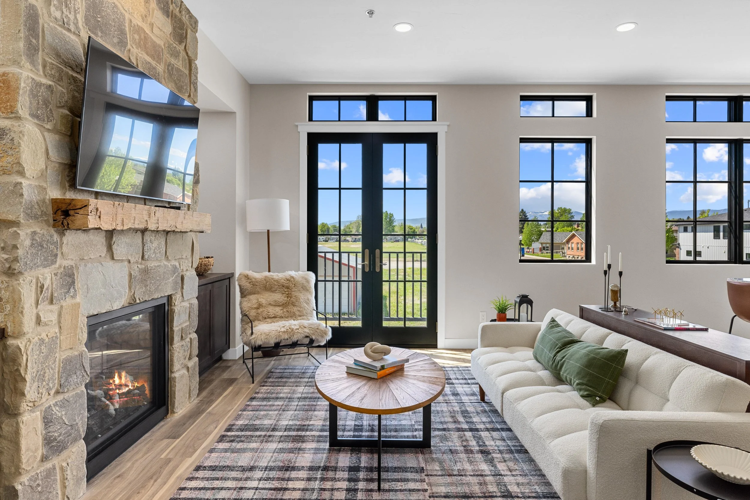 Bright living room with large windows, a white sofa with green pillows, a wooden coffee table on a patterned rug, a stone fireplace with a TV mounted above, a fur-covered chair, and a view of green fields and trees outside.