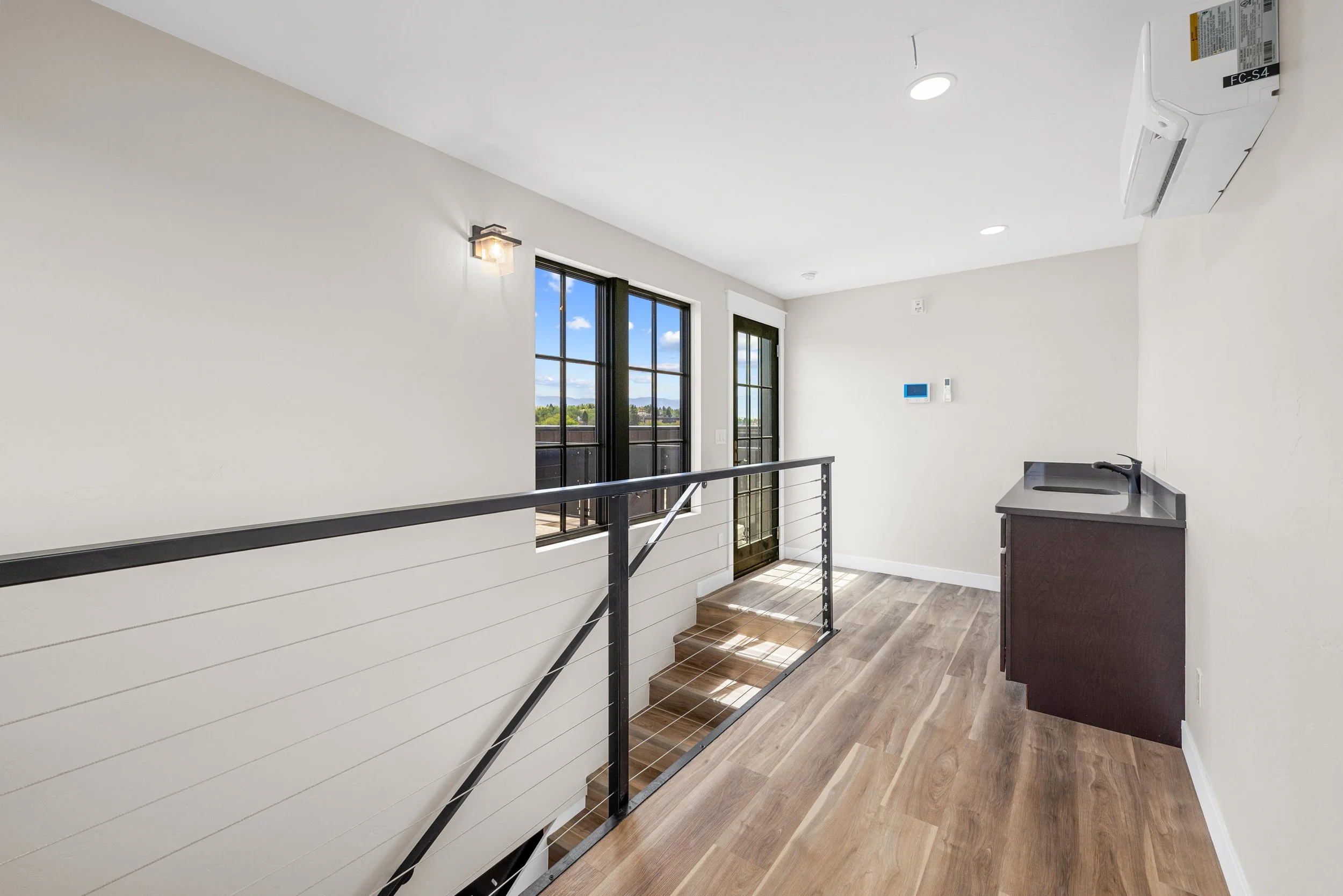 Interior view of a modern apartment with a small kitchenette, wooden floor, railing for stairs, two black-framed glass doors leading to a balcony, and a white wall with a thermostat and an air conditioning unit.