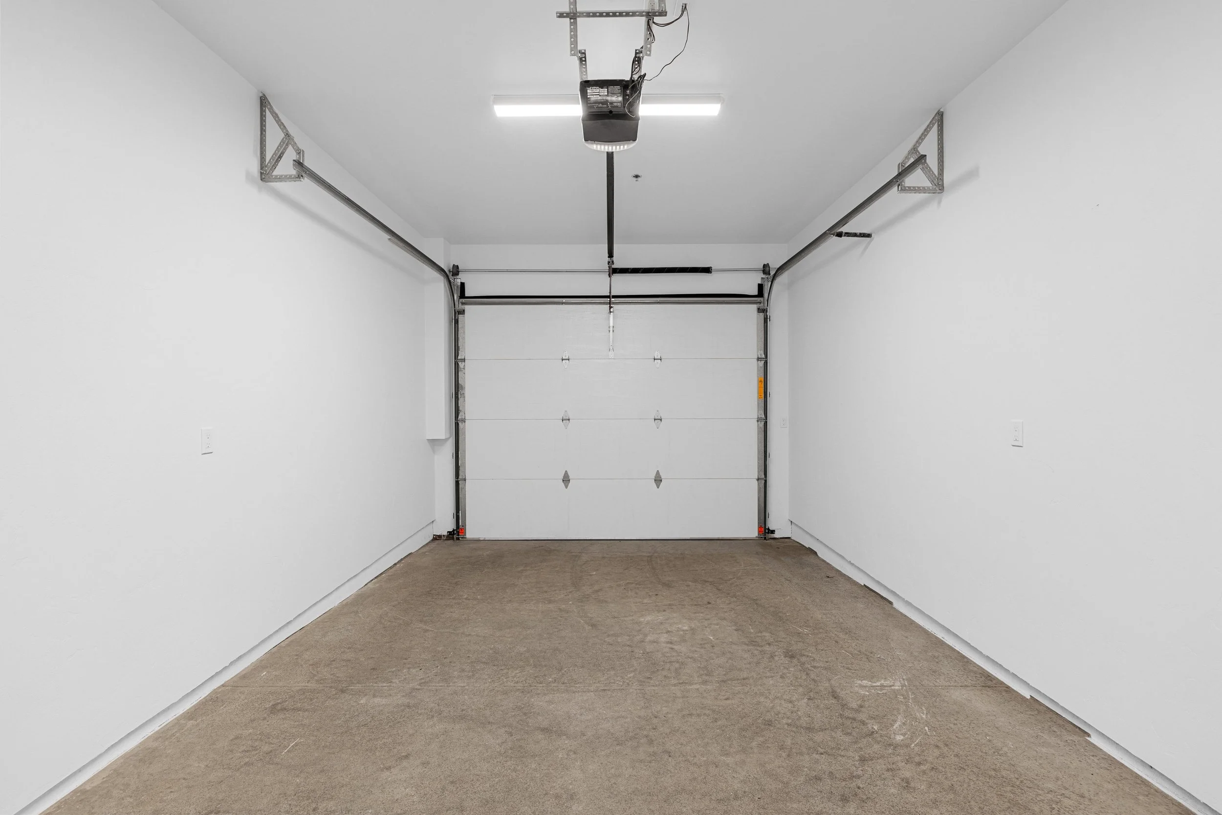 Empty clean garage with white walls, brown concrete floor, and a closed white garage door.