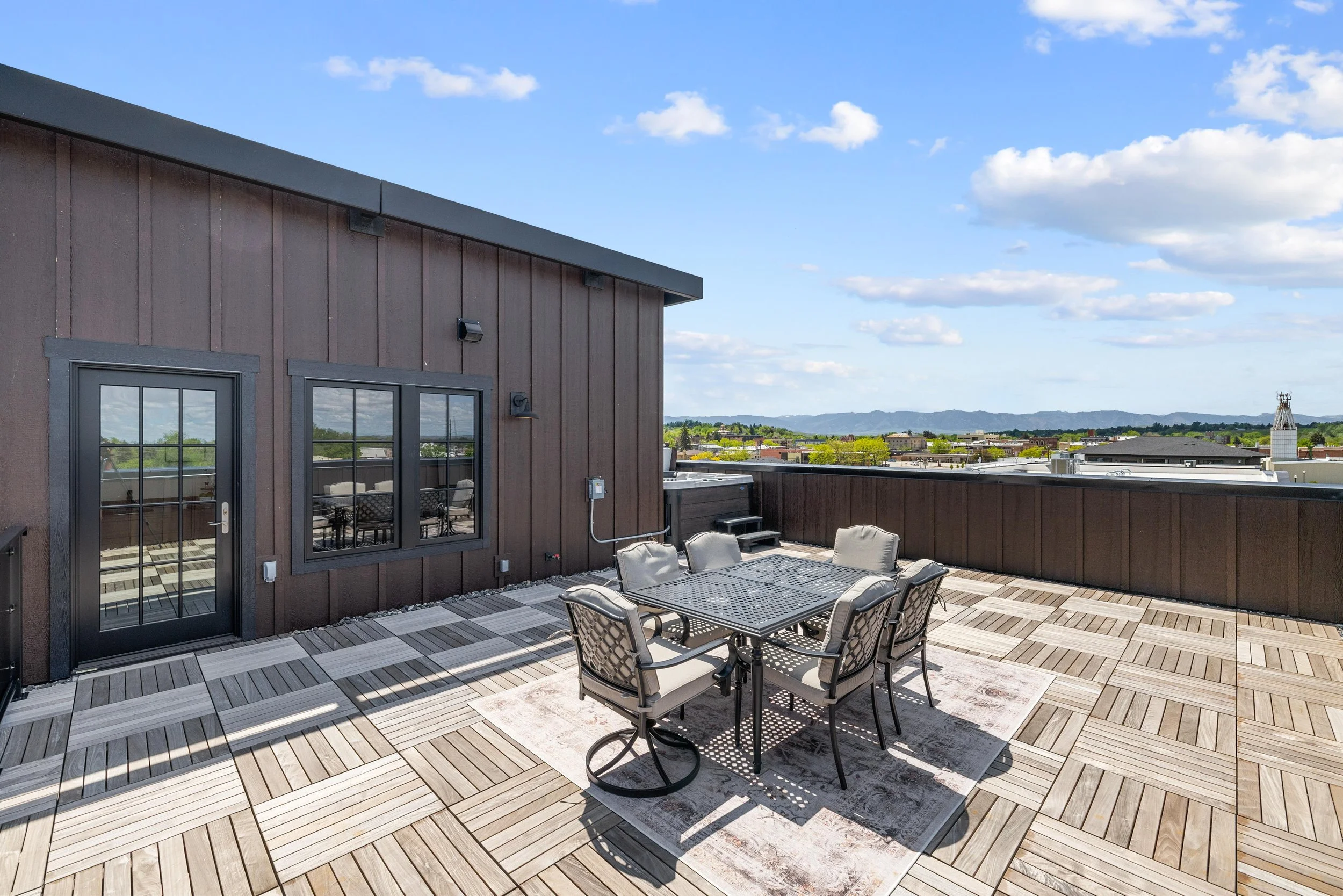 Outdoor rooftop patio with a dining table and six chairs, surrounded by a wooden floor and a brown building wall, with scenic distant hills and cloudy sky in the background.