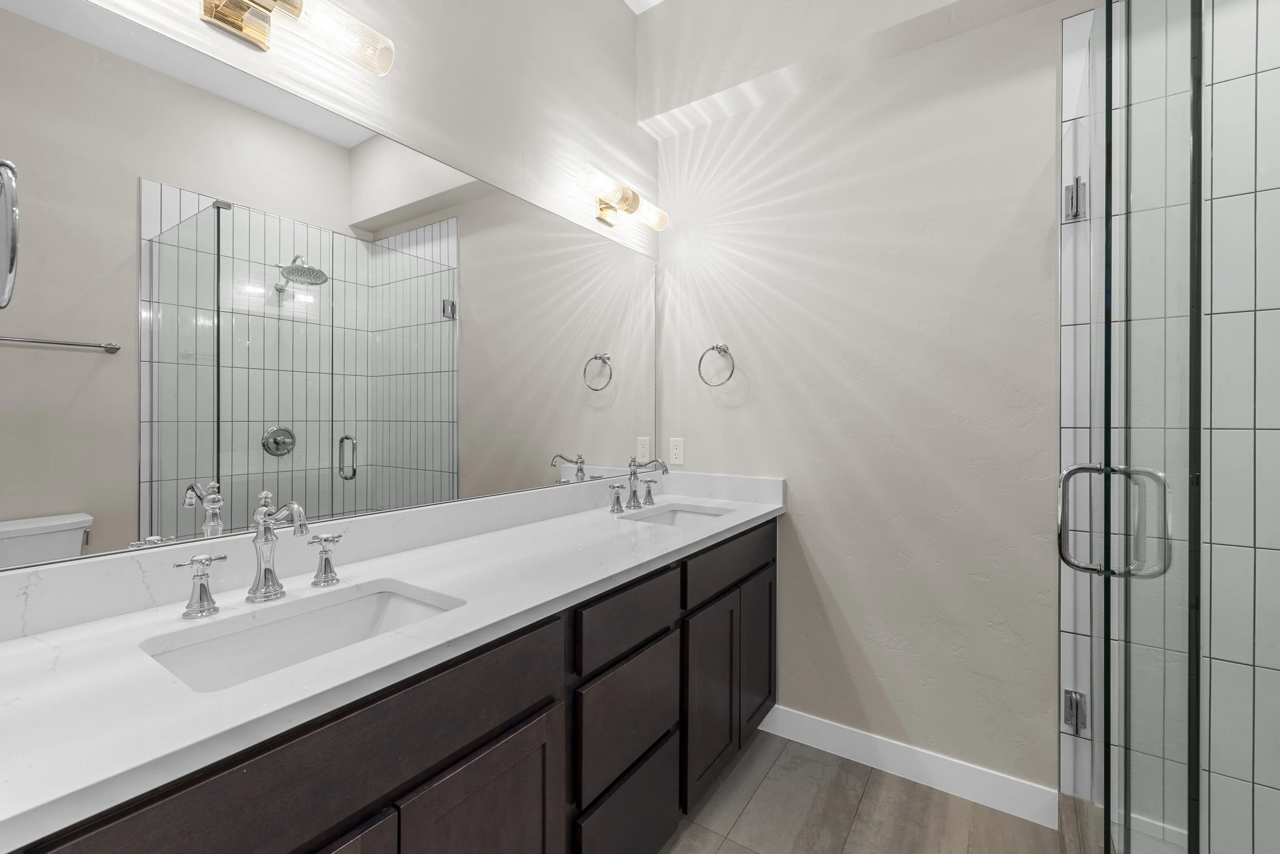 Modern bathroom with a double vanity, dark cabinets, white countertop, large mirror, shower with glass door, and beige walls.