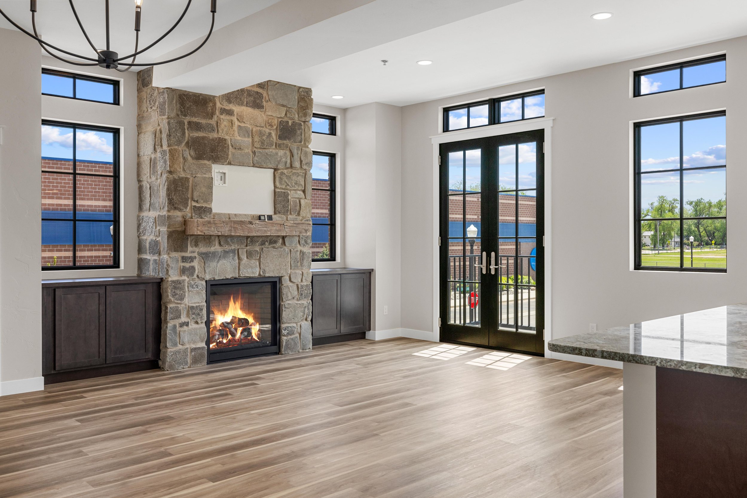 Interior view of a modern living area with a stone fireplace, black window frames, and large windows showing the outside. There are double black doors leading outside, and hardwood flooring.
