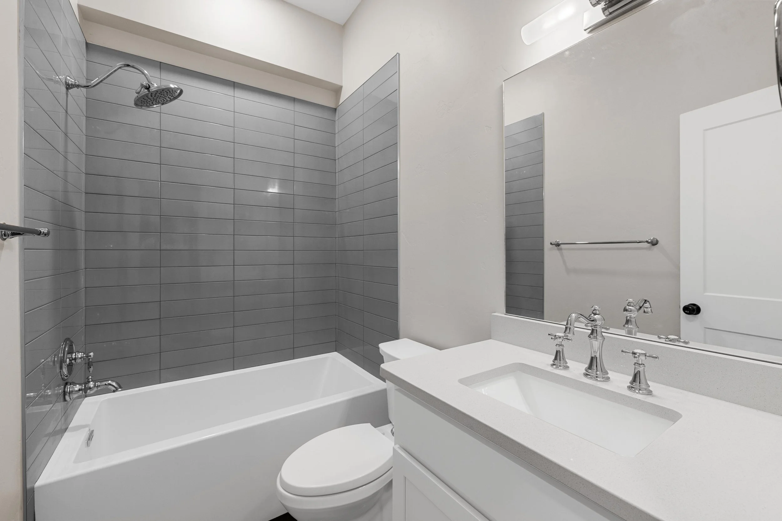 A modern bathroom with a bathtub, a white toilet, a white vanity with a sink, a large mirror, gray tiled walls around the bathtub, and a towel bar on the wall.
