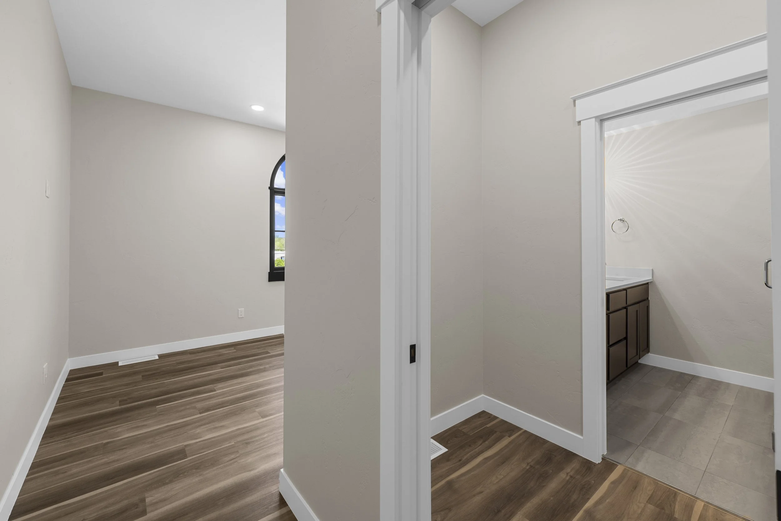 Empty interior room with beige walls, wood-look flooring, arched window, and a small adjacent bathroom with dark vanity, beige tiles, and a mirror
