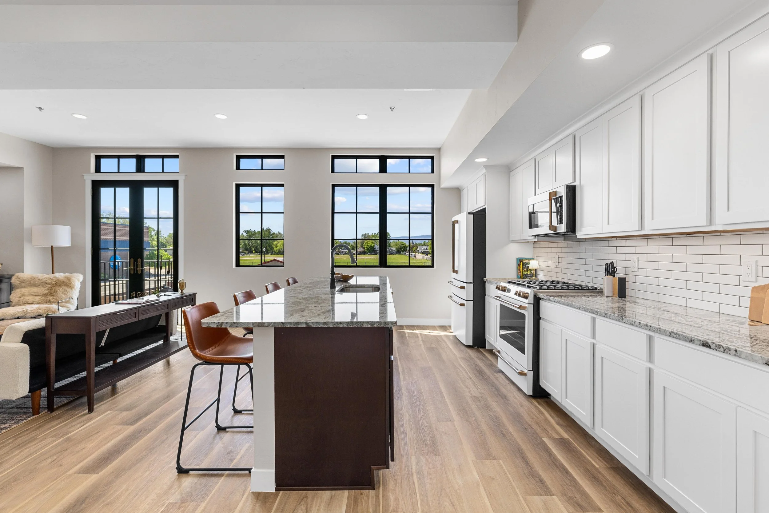 Kitchen with white cabinets, granite countertops, stainless steel appliances, hardwood flooring, and large windows overlooking a green outdoor area.