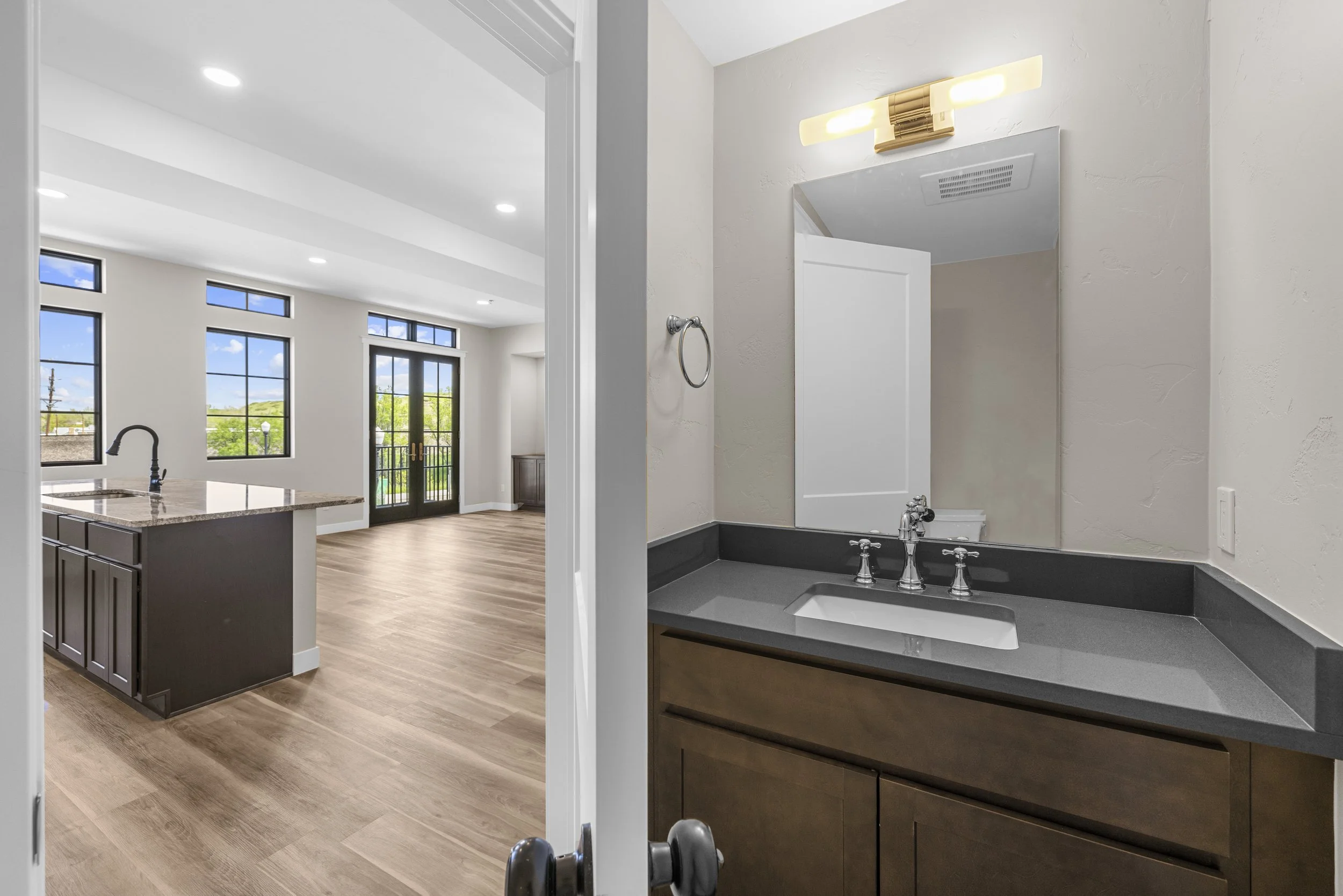 View from a bathroom doorway into an open-concept living room and kitchen with large windows and a balcony door, hardwood floors, and modern fixtures.