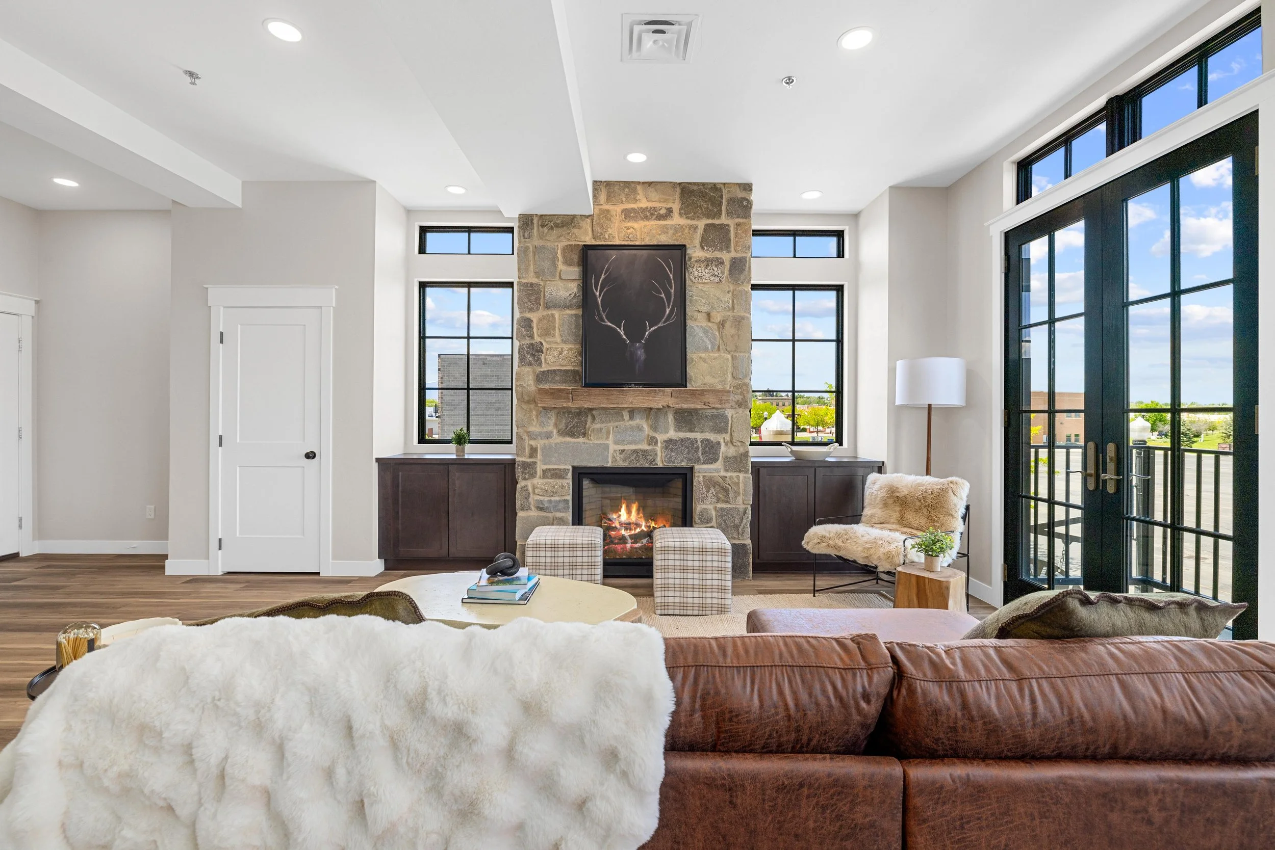 Living room with a stone fireplace, large windows, and a sliding glass door. Furniture includes a brown leather sofa with a white furry throw, a beige armchair with a furry cover, a round coffee table with books, and checked ottomans. Decor features 