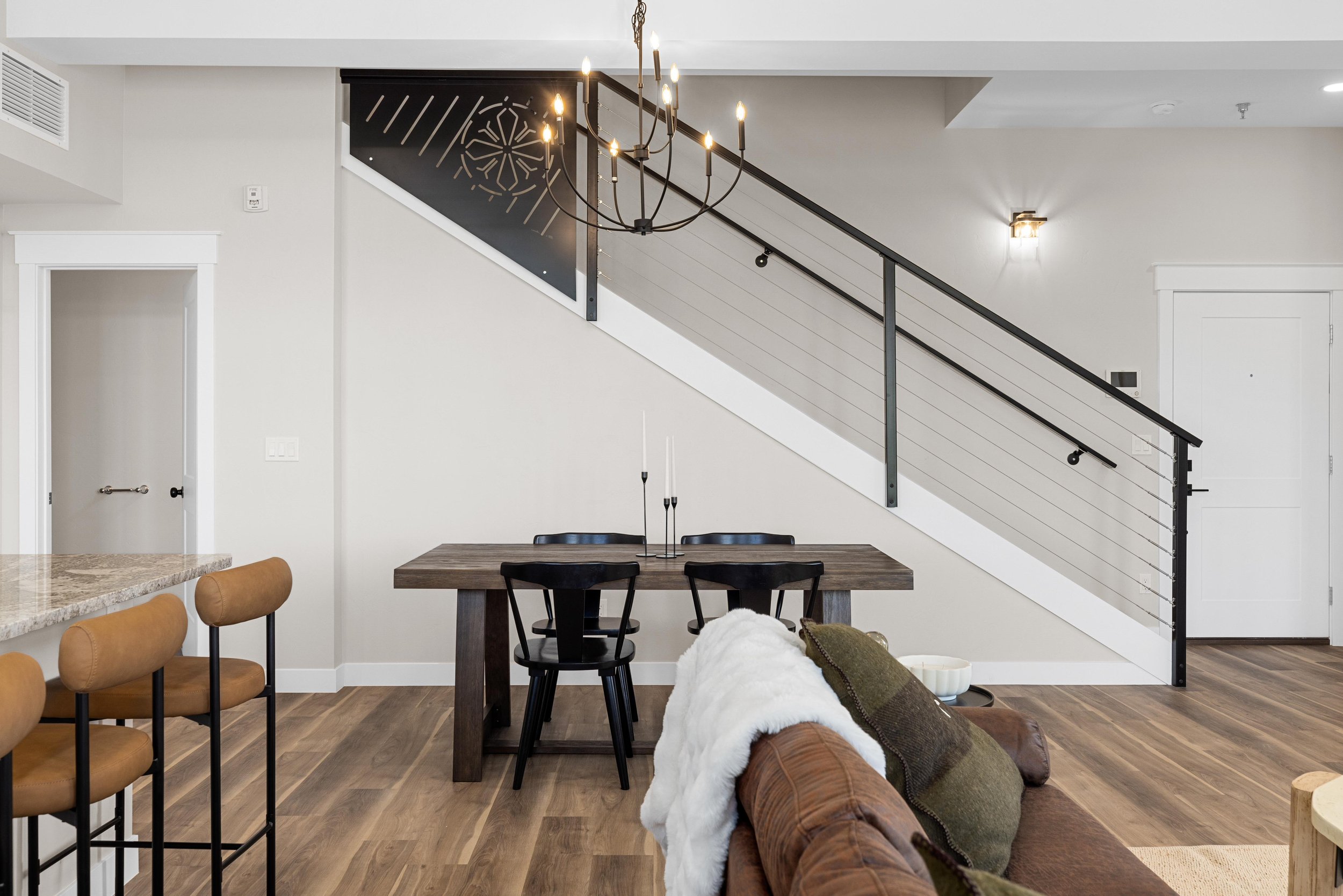 Interior of a modern living and dining area with wood flooring, black and brown furniture, a staircase with black railings, and a chandelier hanging from the ceiling.