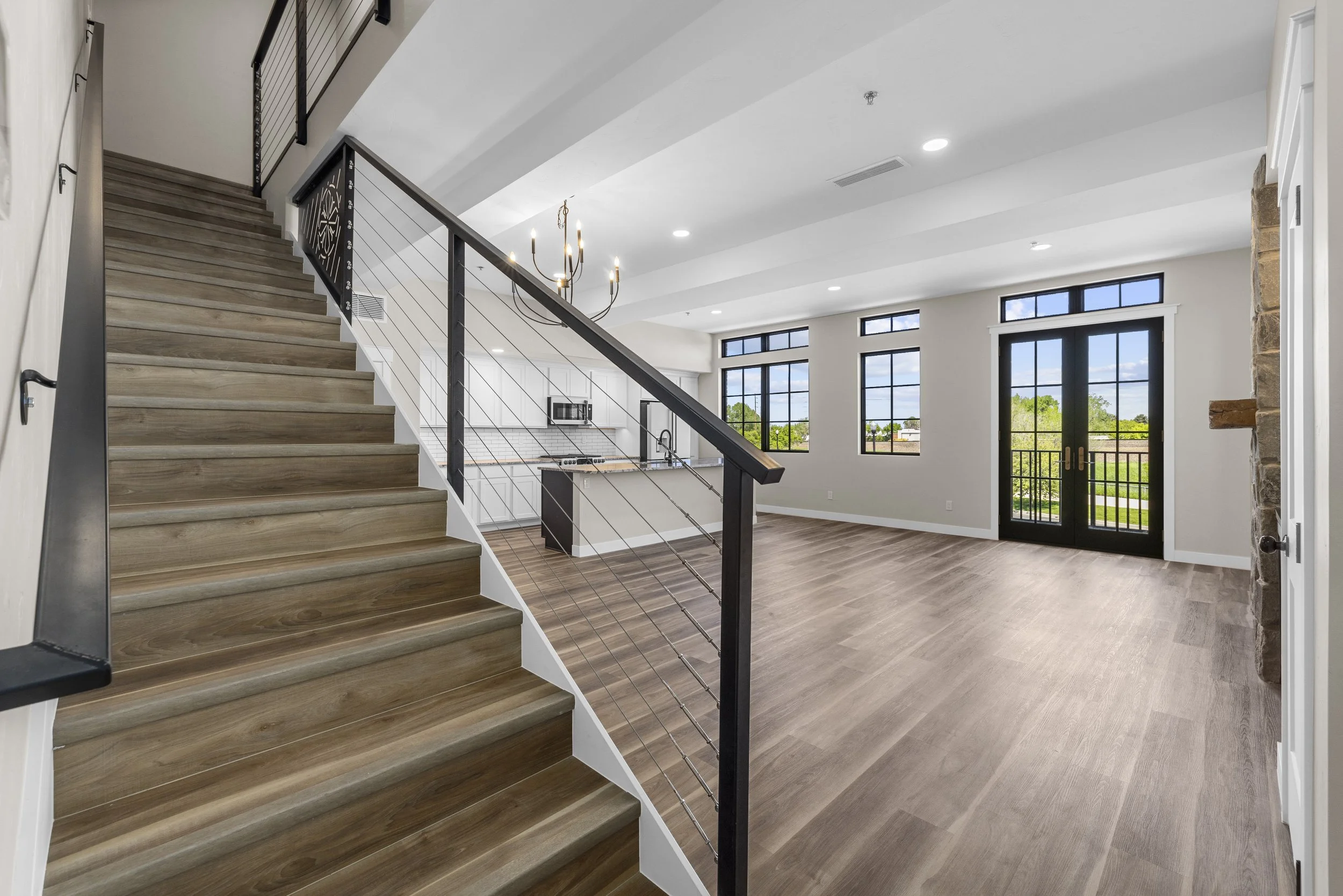 Open-concept living space with wooden flooring, large windows, a kitchen with an island, and a staircase with black metal and cable railing.