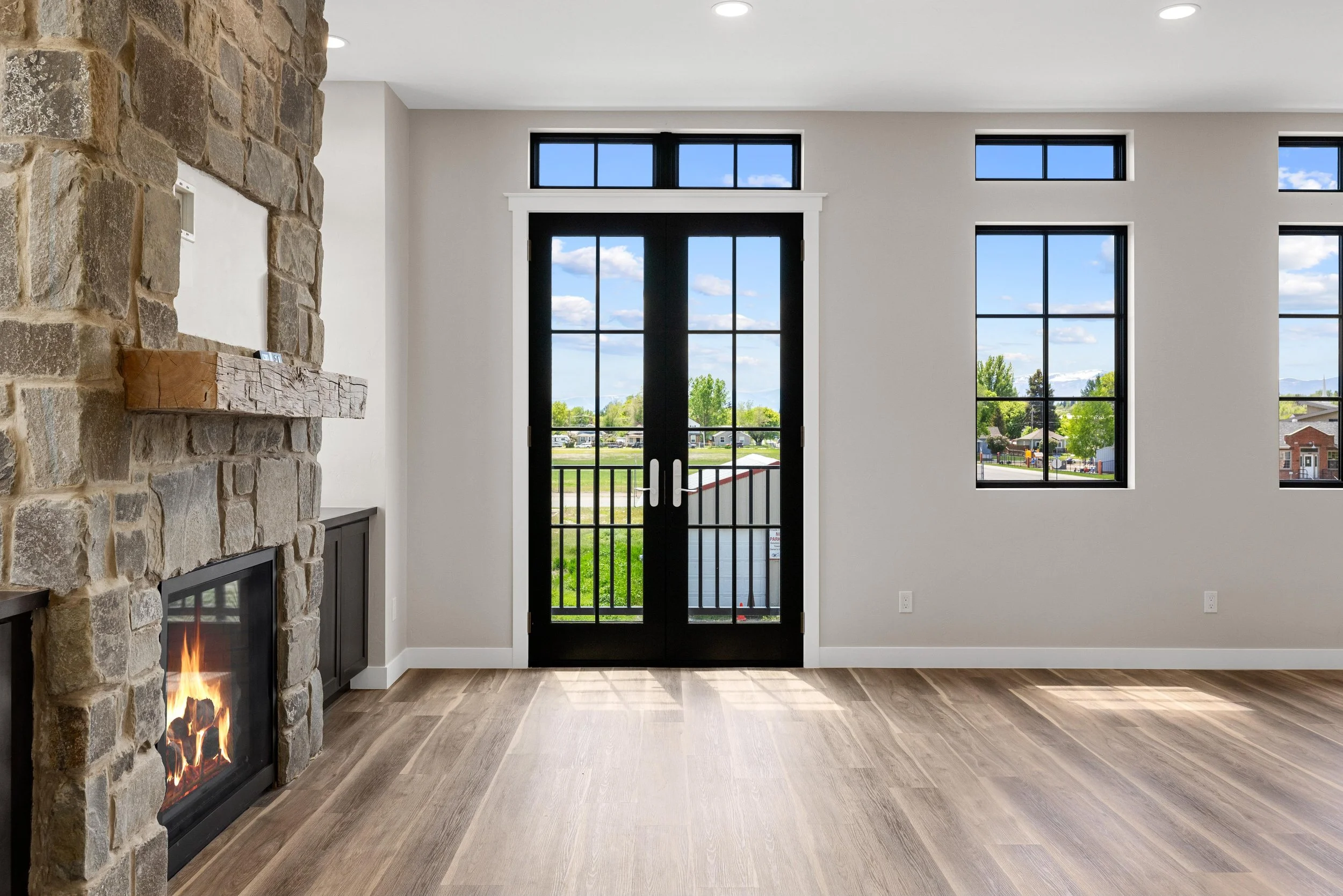 A modern living room with a stone fireplace on the left, large black framed glass doors in the middle, and three large windows on the right showing a view of greenery and houses. The room has light-colored walls, wooden flooring, and a minimalist sty