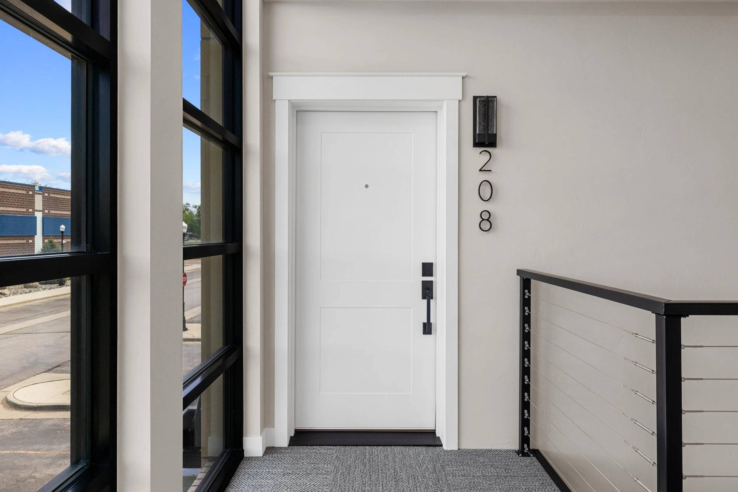 Interior view of a modern apartment or office building entrance door, with black metal and glass windows on the left, a white door with black handle and a small peephole in the center, a black wall-mounted light fixture, and black metal numbers '208'