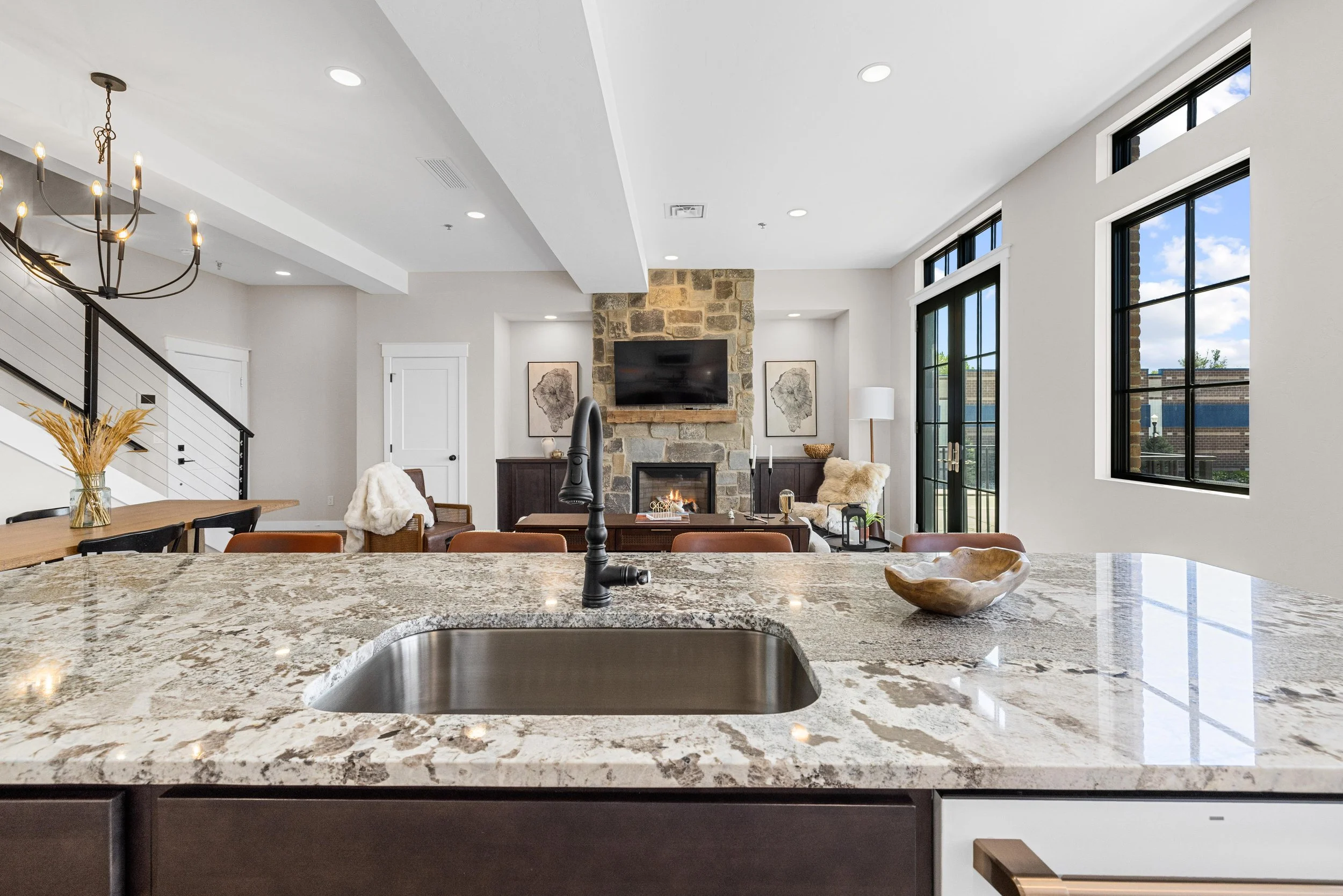 Modern open-concept living room seen from kitchen with granite countertop in foreground, stone fireplace, wall-mounted TV, large windows, and furniture with decor.