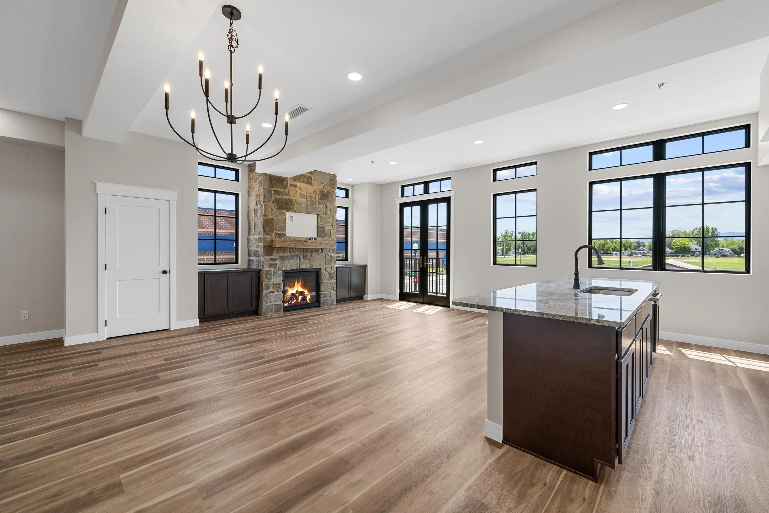 Modern open-concept living space with hardwood floors, a stone fireplace, large windows, a chandelier, and a kitchen island with a sink.