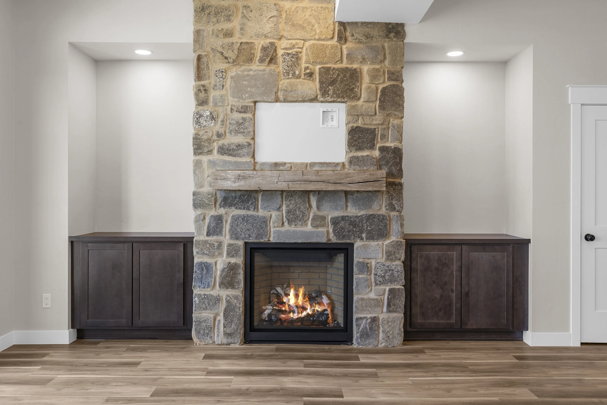 Living room with a stone fireplace, two dark wood cabinets on each side, wooden mantel, and a fire burning inside.