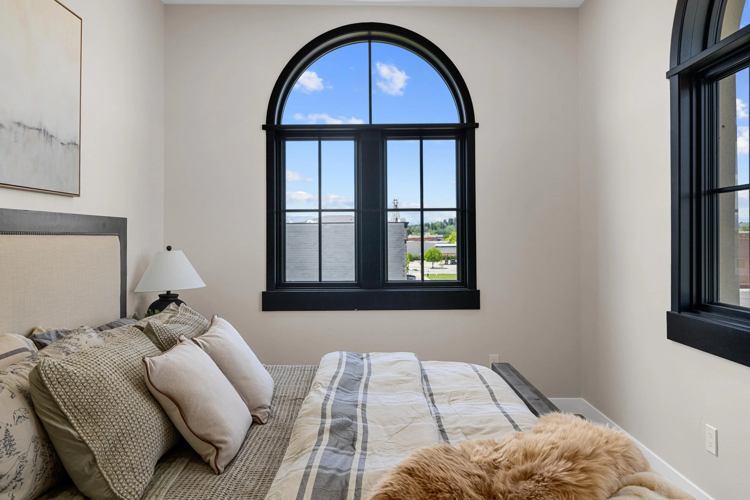 An Ice Haus Condo Bedroom with a beige bed, multiple pillows, light-colored blanket, black-framed arched and rectangular windows showing a view of a cloudy sky and buildings outside.
