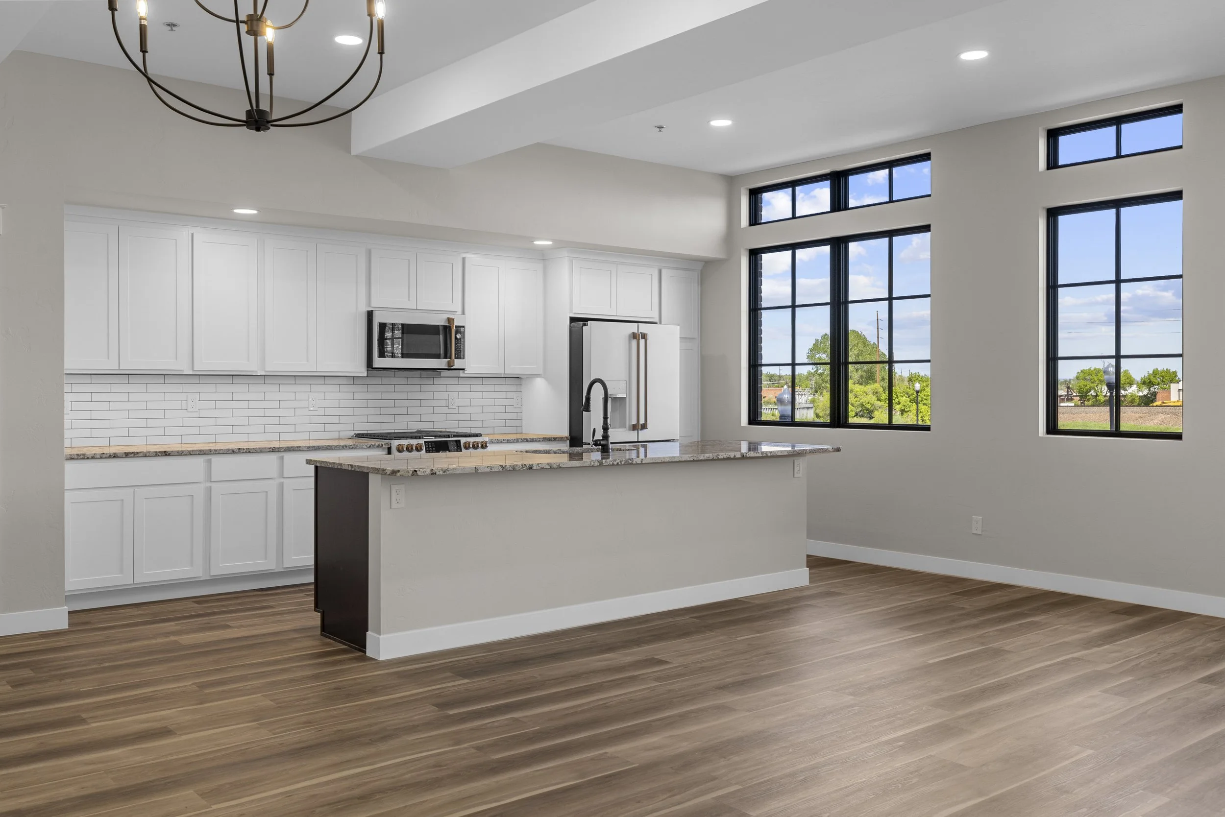 Empty modern kitchen with white cabinets, granite countertops, white subway tile backsplash, stainless steel microwave, and refrigerator. Large windows overlooking green trees, hardwood floors, and a black chandelier hanging from the ceiling.