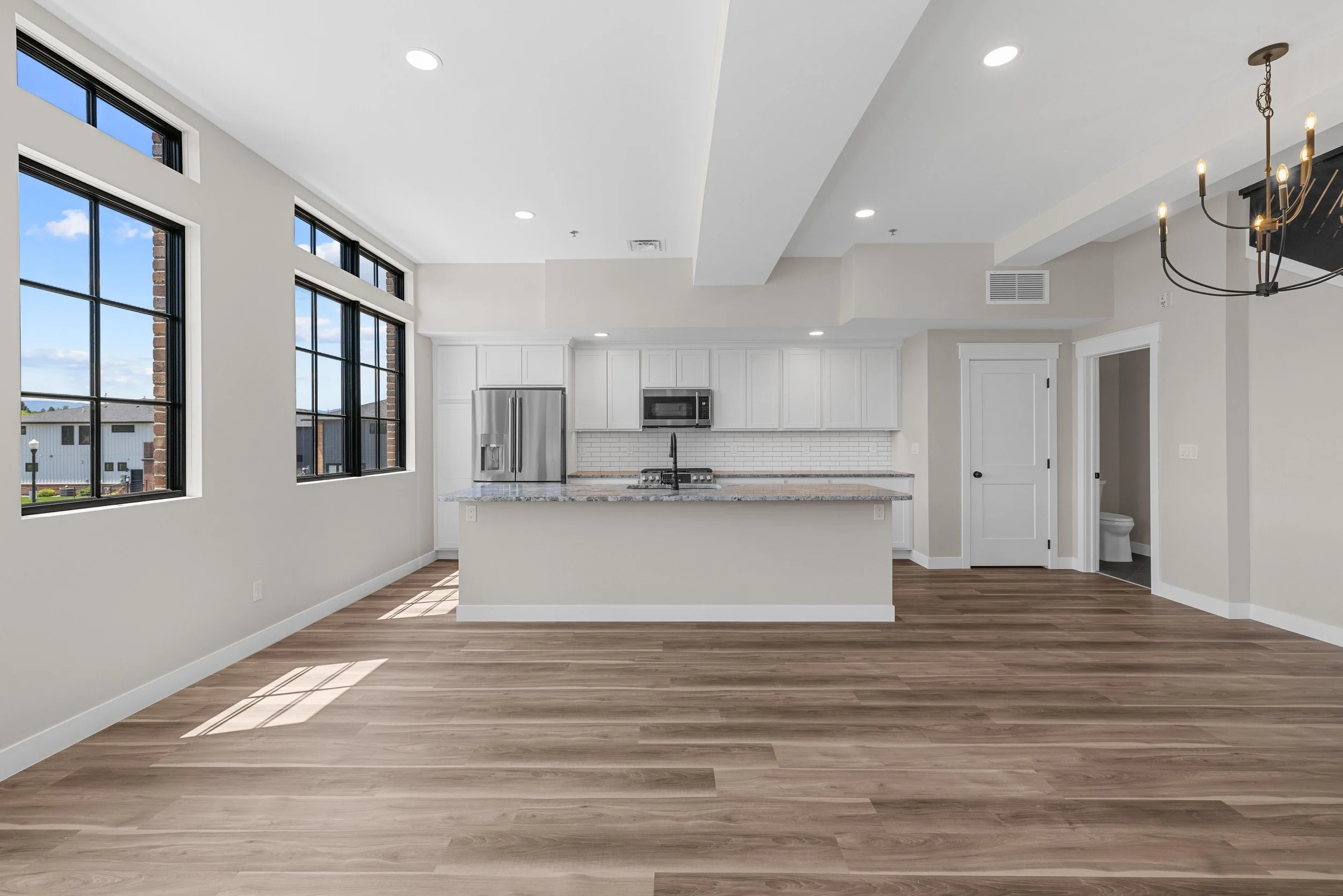 Modern kitchen with white cabinets, stainless steel refrigerator, granite island, wood flooring, and large black-framed windows.