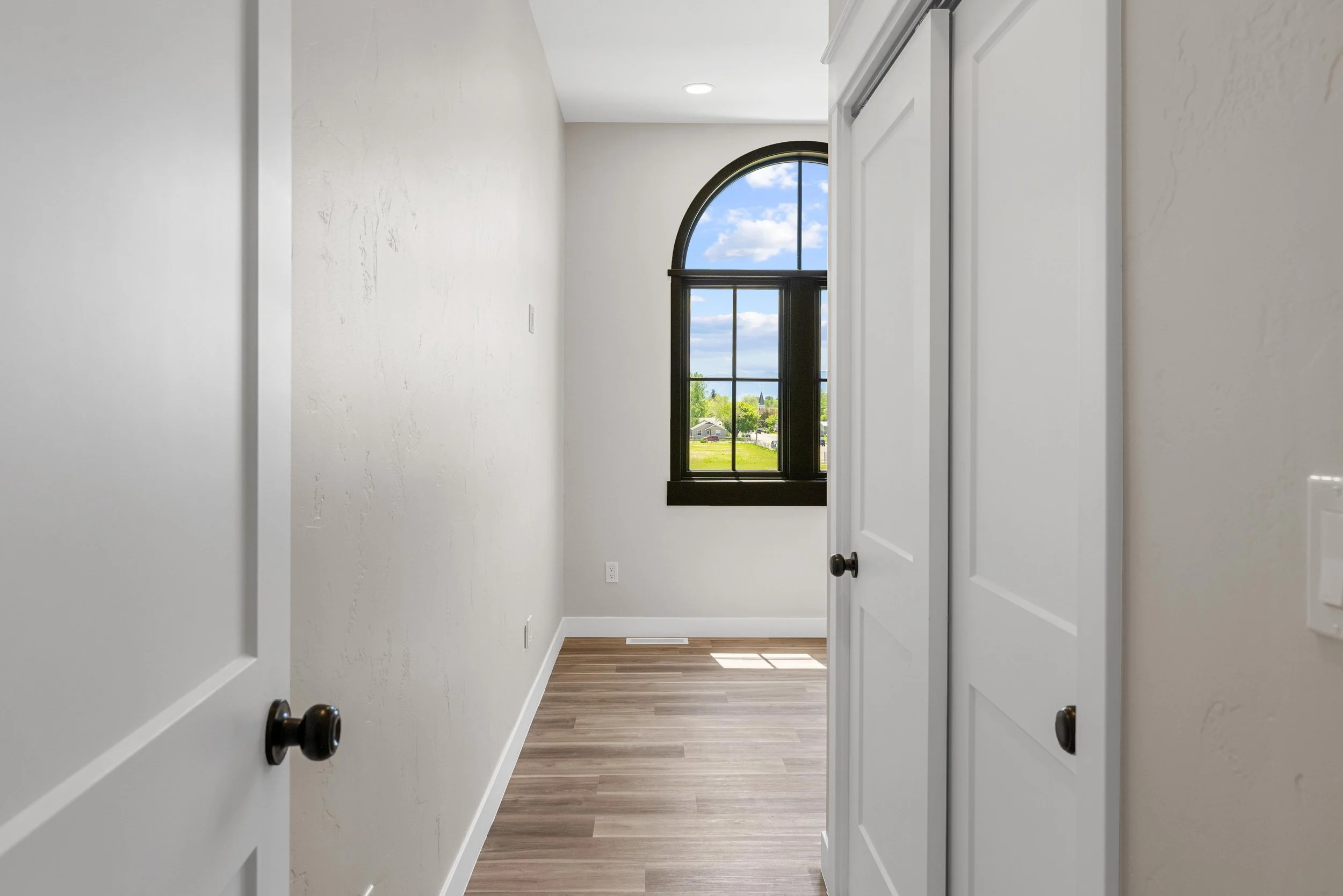 Empty room in an Ice Haus Condo for sale in Sheridan, Wyoming,  with white walls, a hardwood floor, black window frame, and a large arched window showing a sunny outdoor scene.
