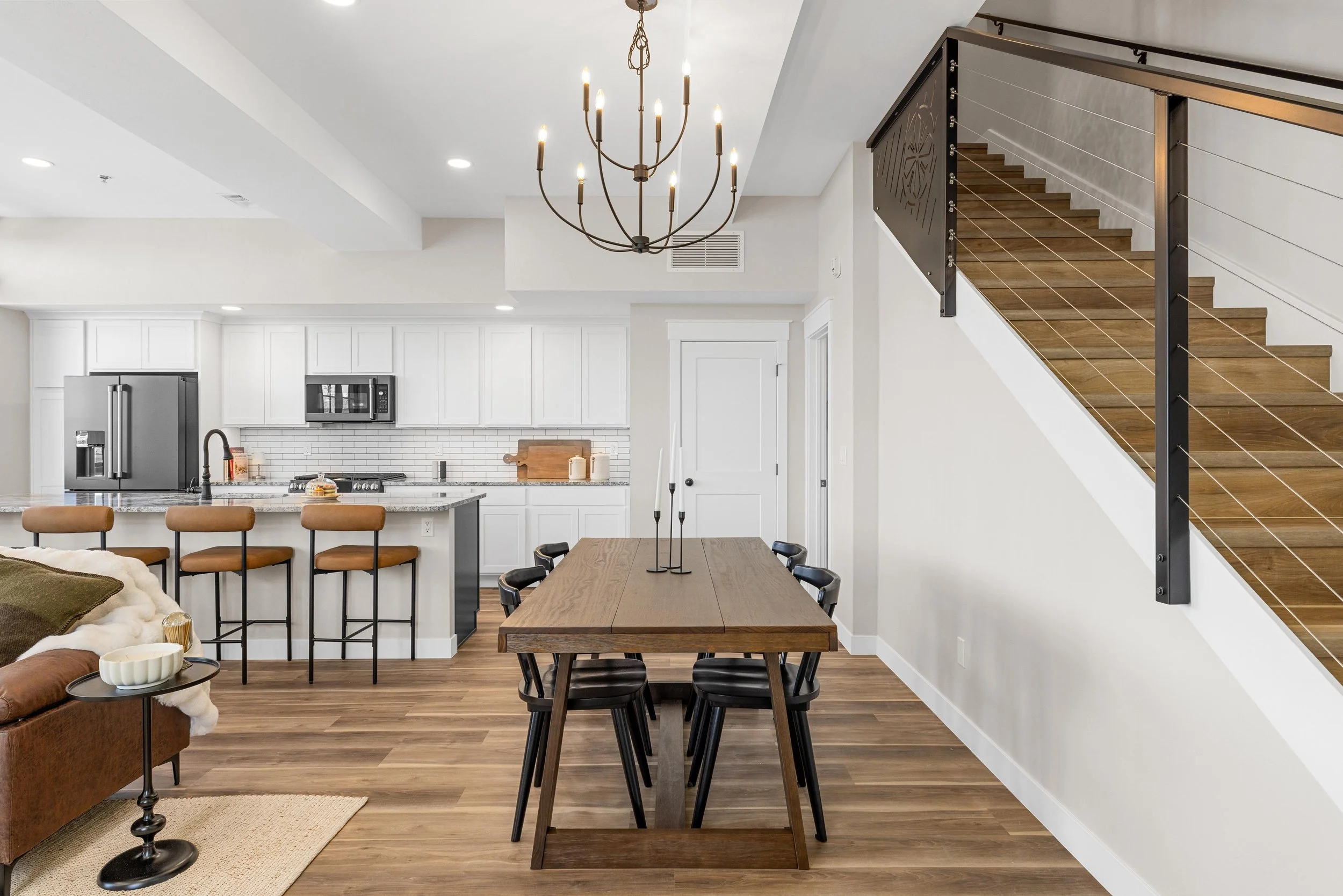 Open-concept kitchen and dining area with wooden flooring, white cabinets, stainless steel appliances, a wooden dining table with black chairs, and a staircase with wood steps and black metal railing. A chandelier hangs from the ceiling.