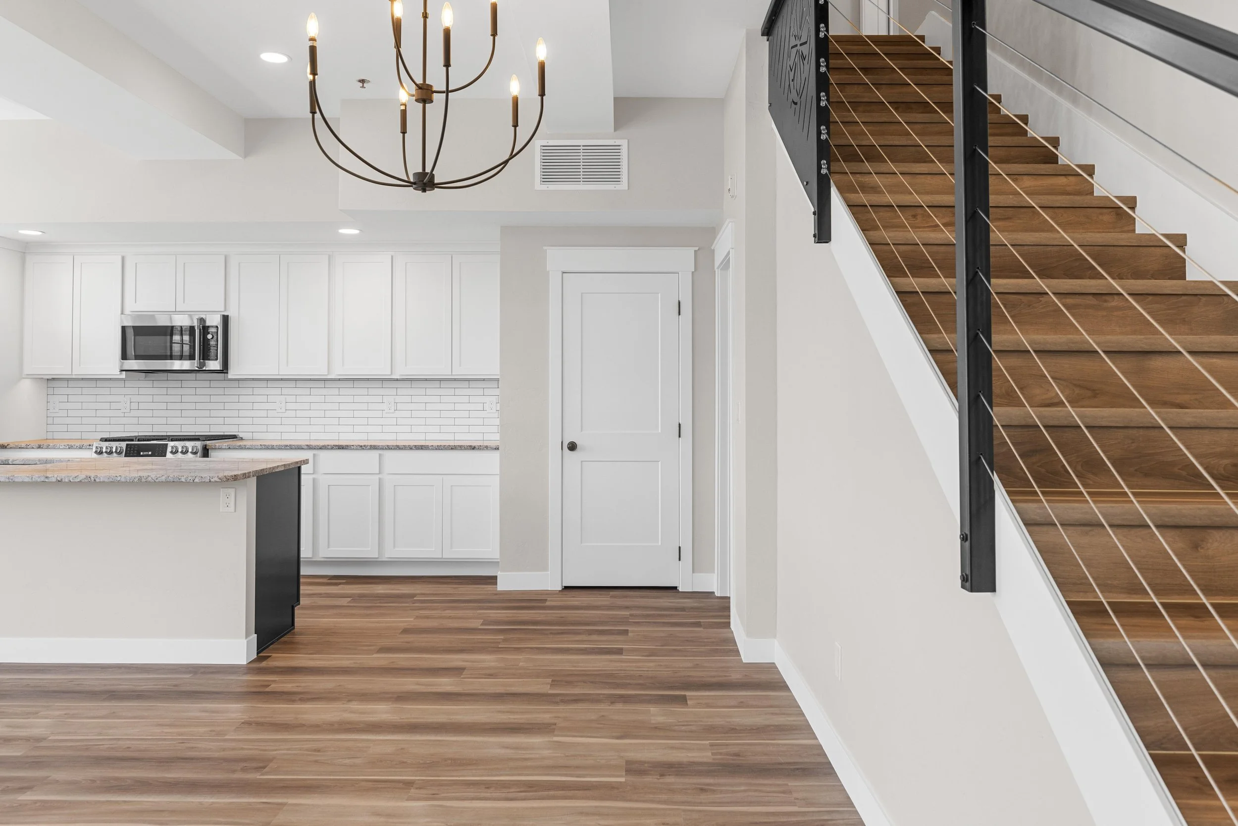 Interior view of a modern kitchen and staircase, featuring white cabinets, a brick backsplash, wooden flooring, and a black staircase railing with a chandelier overhead.