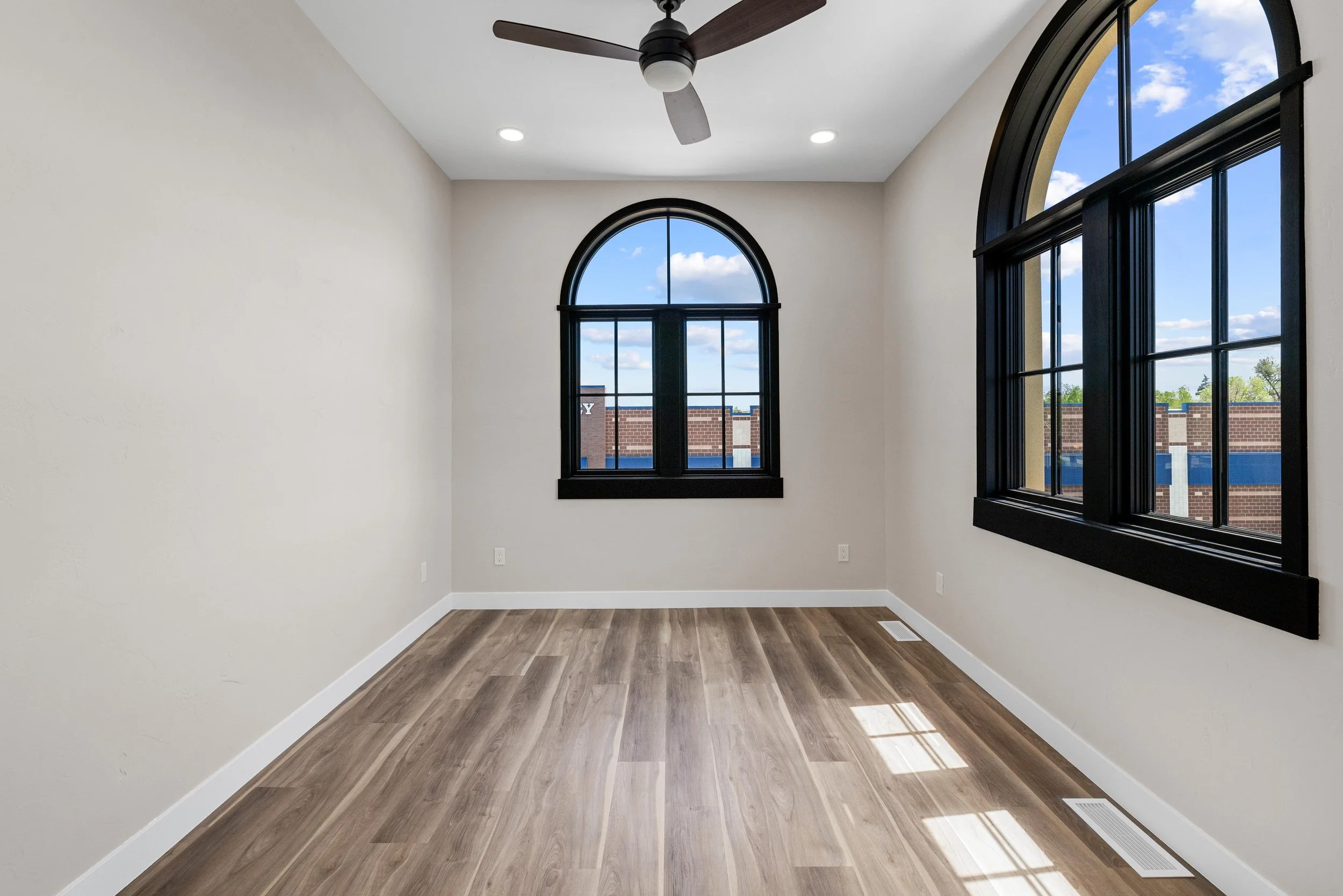 An empty room with cream-colored walls, hardwood floors, black window frames, a ceiling fan, and natural light coming through large arched windows.