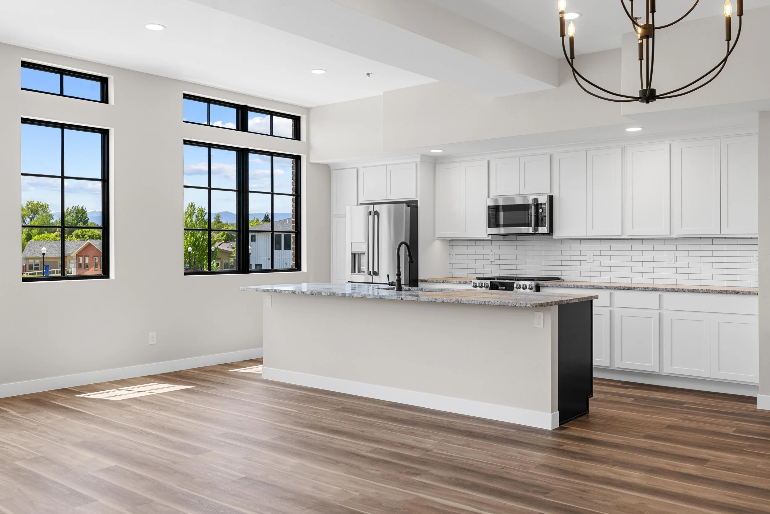 Empty modern kitchen with white cabinets, stainless steel refrigerator, microwave, and stove, large windows, wooden floor, and a black chandelier.