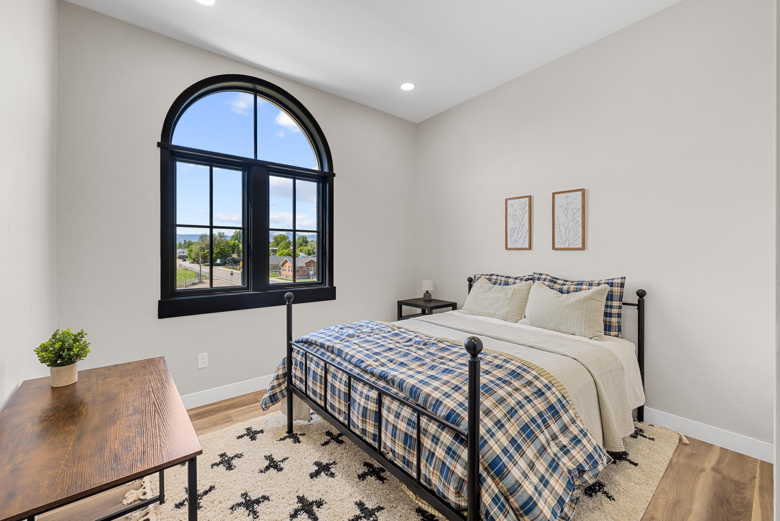 A bedroom with a black metal bed frame, plaid bedding, two pillows, a nightstand with a lamp, framed artwork, a window showing a neighborhood view, and a wooden dresser with a potted plant.