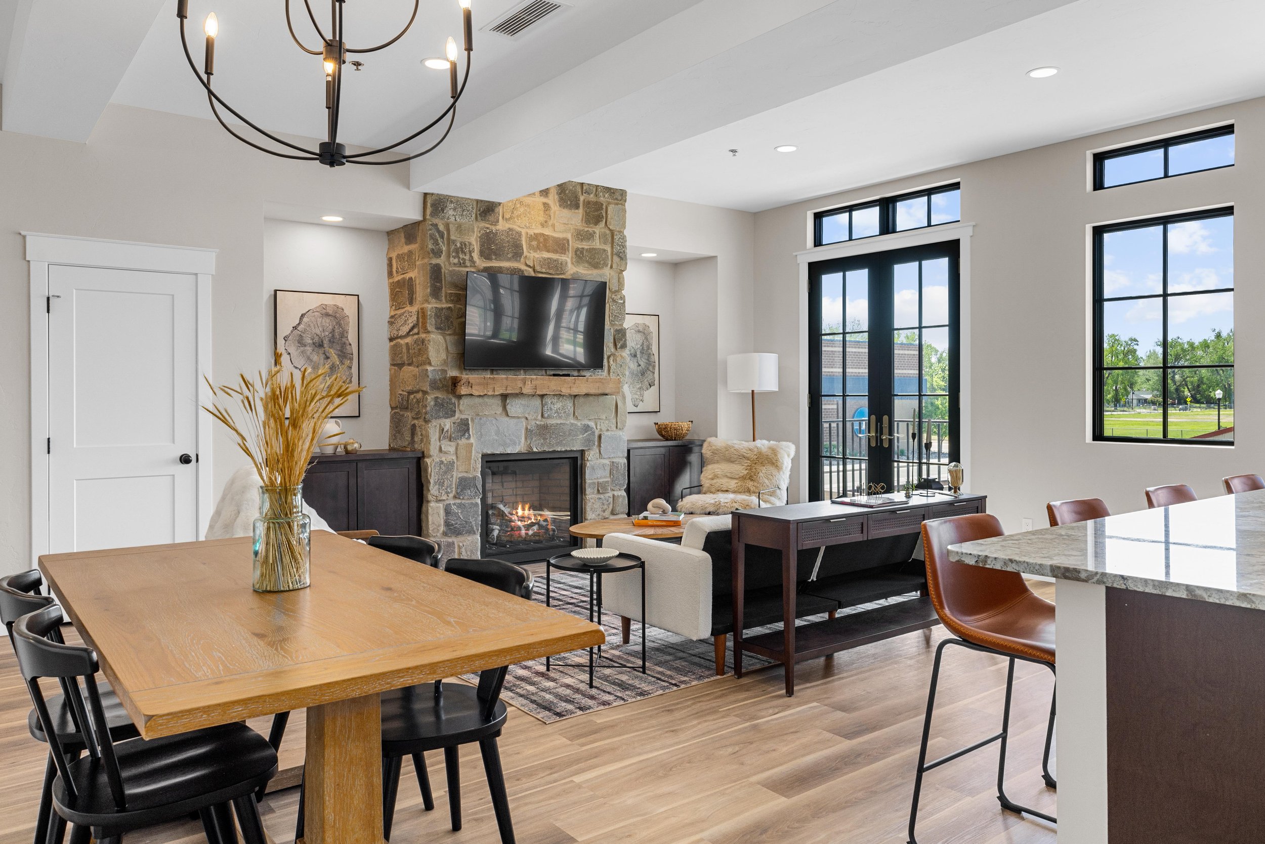 Living room inside of a condo for sale in Sheridan, Wyoming. With a stone fireplace, mounted TV, modern furniture, large windows, and a dining area with a wooden table and black chairs, bright natural light.