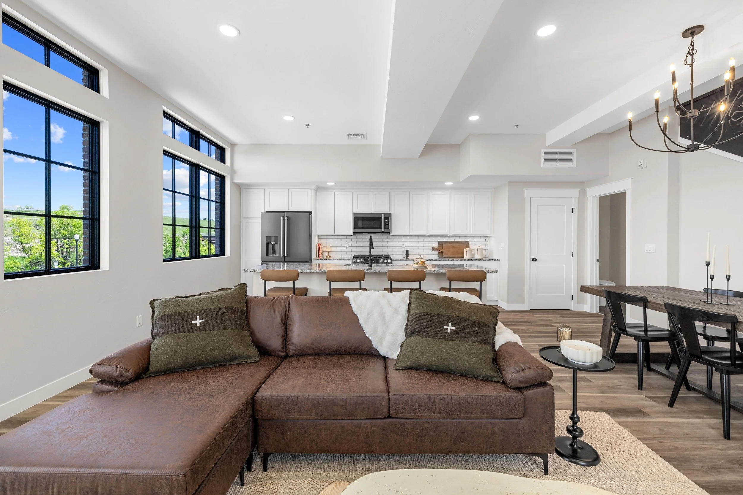 Open-concept living room and kitchen with large windows, brown sectional sofa with pillows, dark wood dining table with candlesticks, and white cabinets in the kitchen.