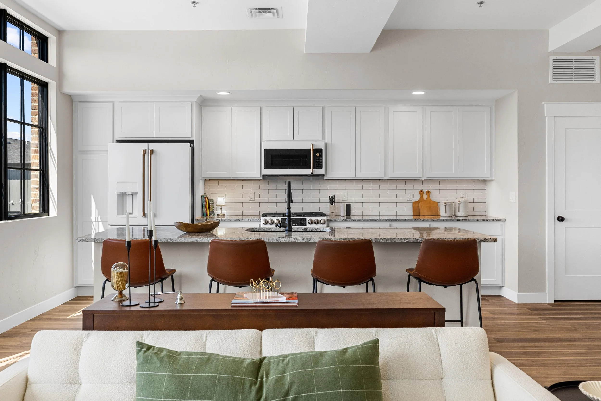 Open-concept kitchen inside an Ice Haus Condo for sale in Sheridan, Wyoming featuring white cabinetry, granite countertops, subway tile backsplash, and island seating.