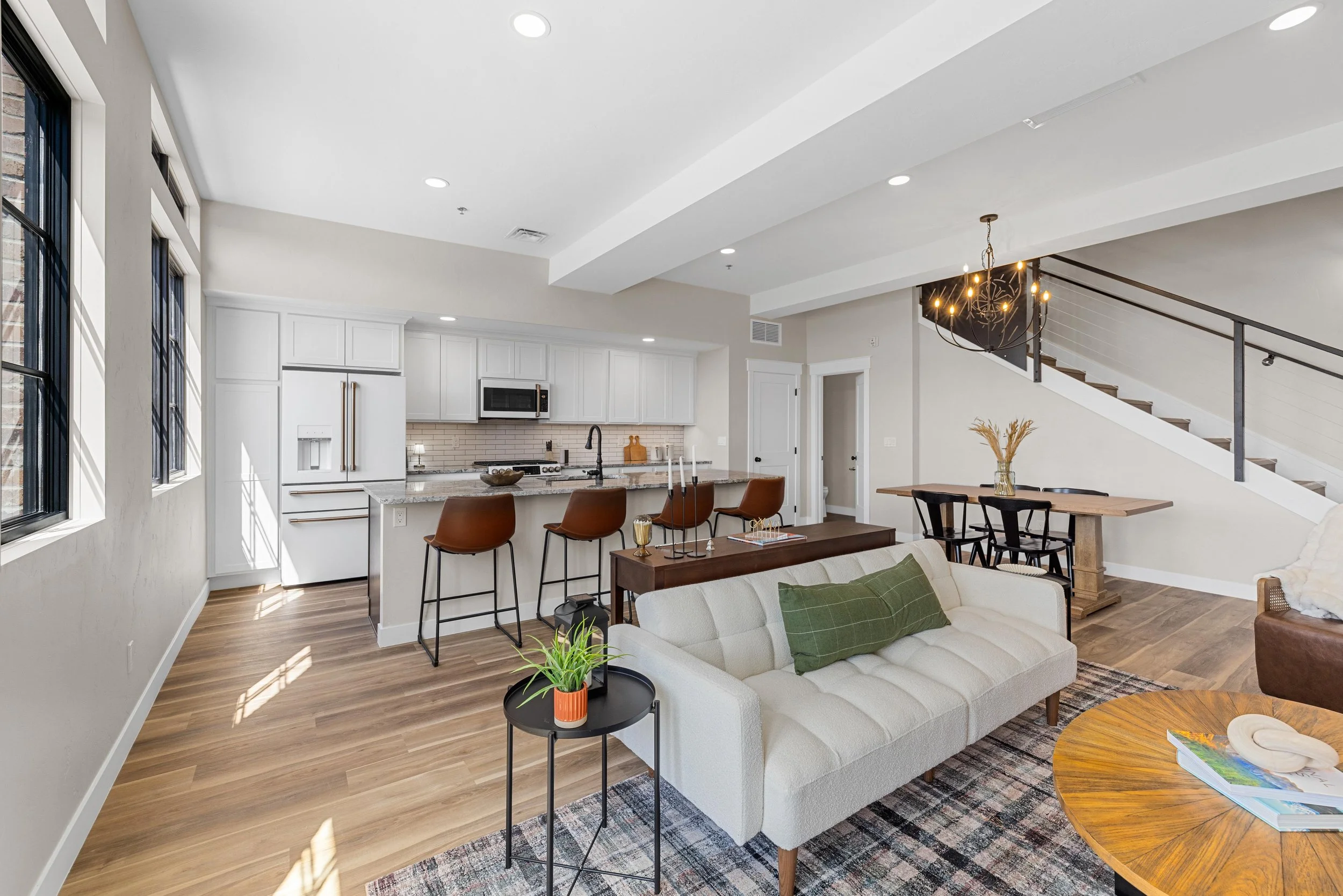 Open-concept living room and kitchen inside an Ice Haus Condo for sale in Sheridan, WY with granite countertops, island seating, dining area, staircase, and modern finishes.